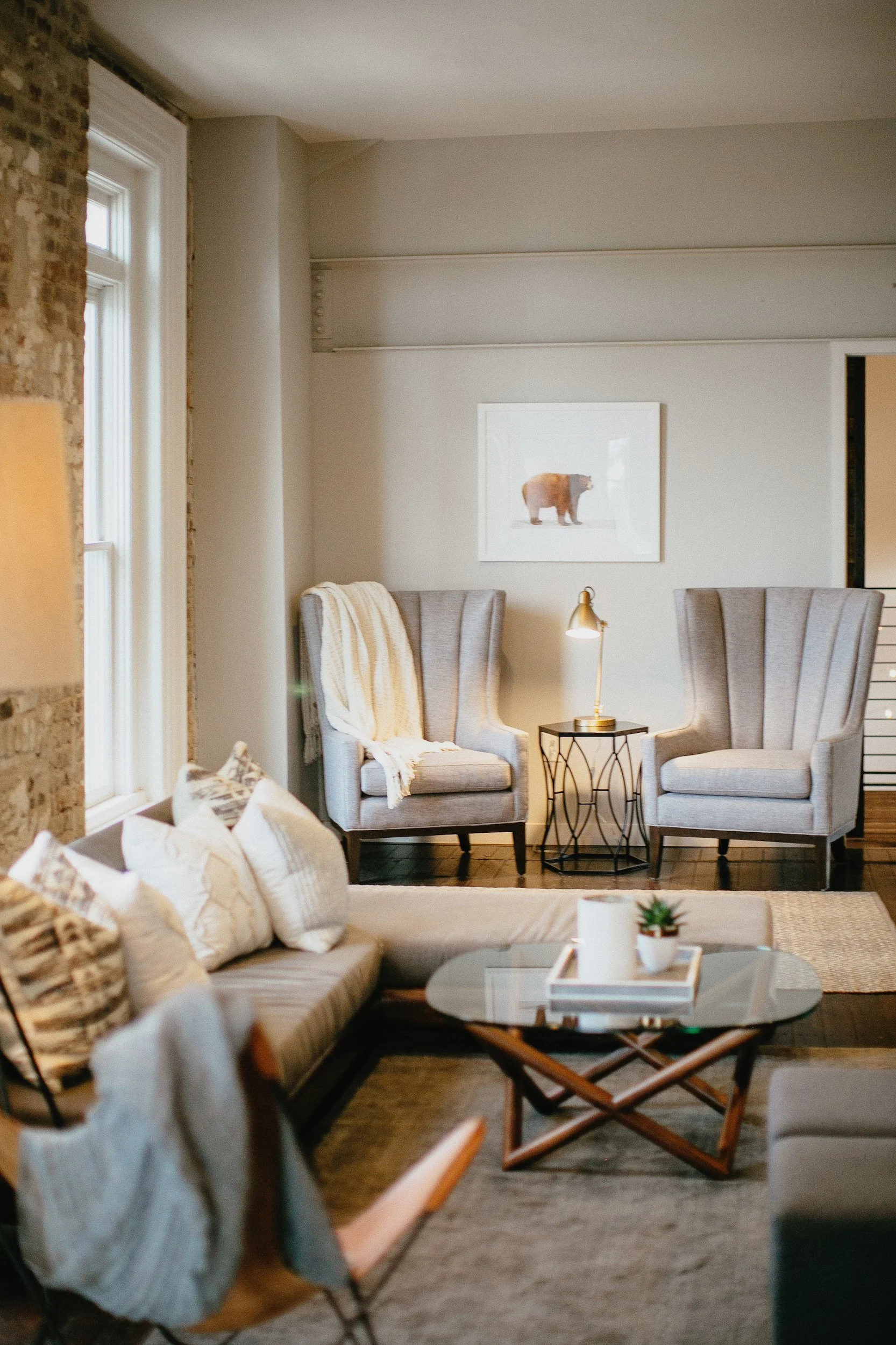 The Loft at the Cordelle private suite and Living room with two gray upholstered armchairs, a sofa with pillows, a coffee table with a tray and plant, and a large window bringing natural light.
