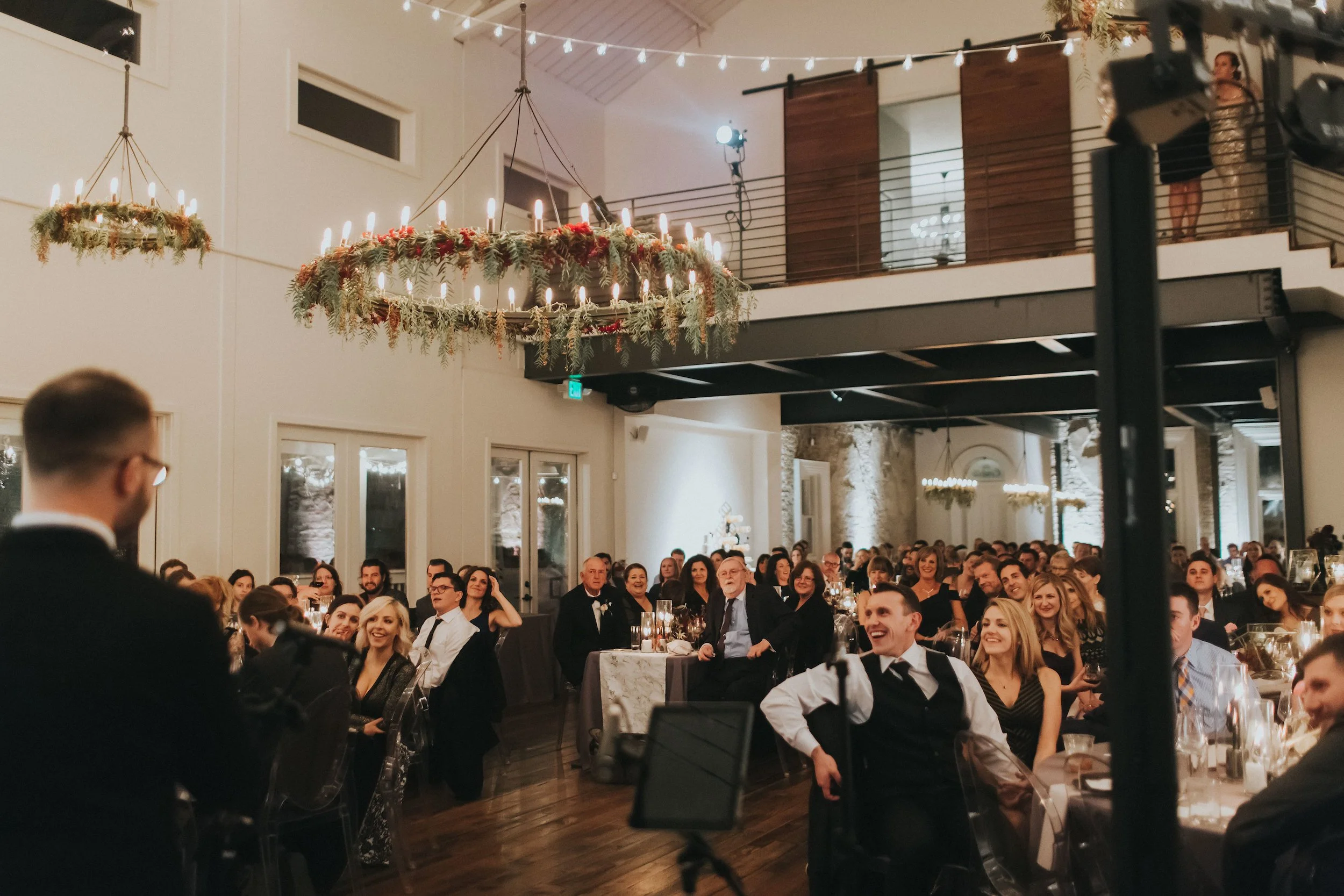 A large group of people dressed in formal attire sitting at tables in a decorated indoor venue for a celebration, with a chandelier decorated with greenery and candles hanging from the ceiling, and string lights overhead.