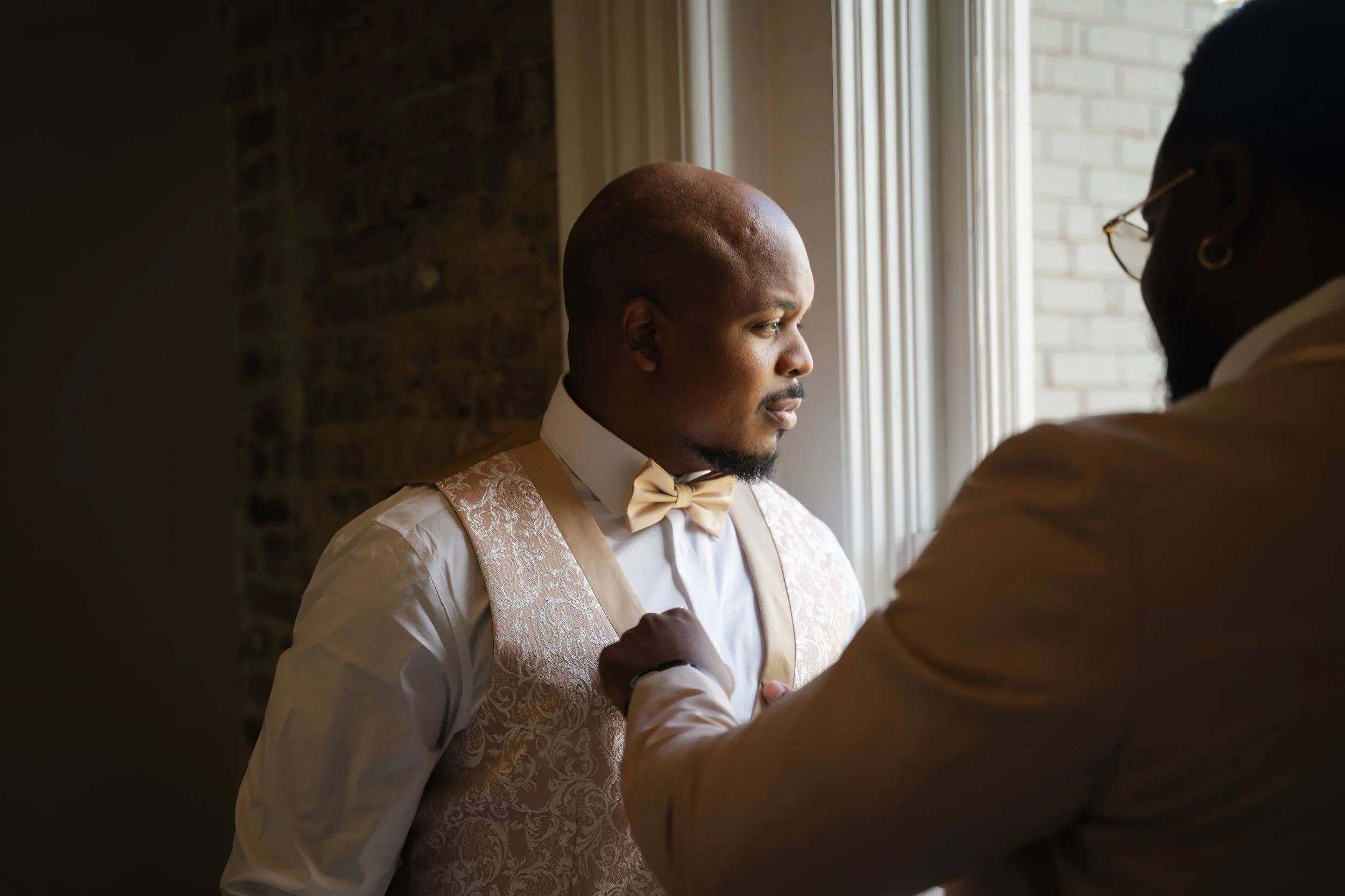 Groom getting ready in the well appointed loft at the Cordelle A man dressed in formal wear, including a patterned vest, white shirt, and bow tie, standing near a window with natural light, while another person adjusts his sleeve or cuff.