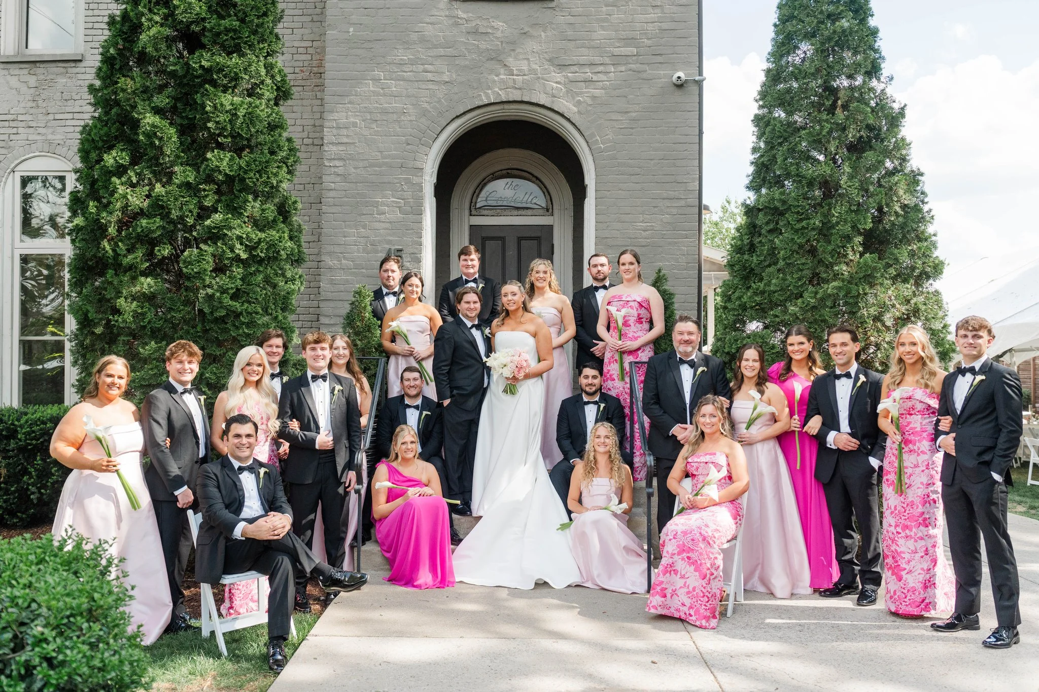 A wedding party in front of the Cordelle in Downtown Nashville in wedding attire, including a bride in a white gown holding a bouquet, and bridesmaids in pink dresses with gray brick walls and tall trees.