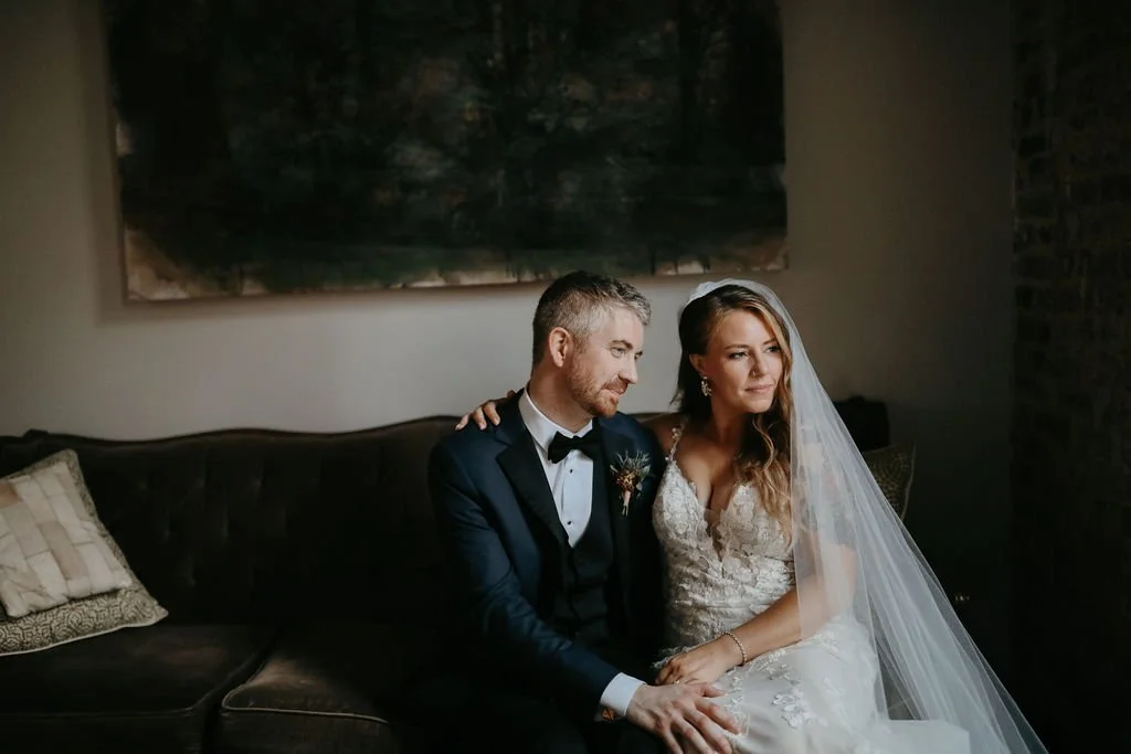 A Cordelle bride and groom sit on a dark brown couch inside, with a large painting or picture hanging on the wall behind them. The groom wears a dark tuxedo with a bow tie, and the bride wears a white lace wedding gown with a long veil.