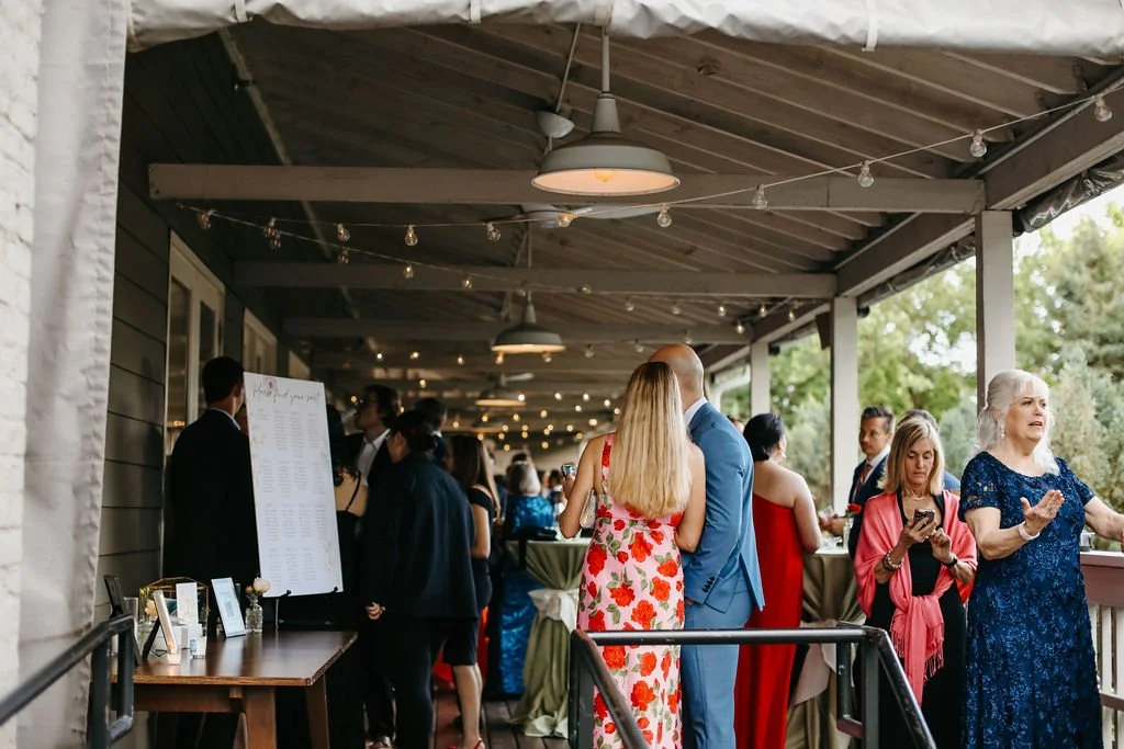 People attending a social gathering or wedding reception on a covered outdoor patio with string lights, some talking and others using their phones. Covered porch for cocktail hour signature cocktails and wedding inspirations.
