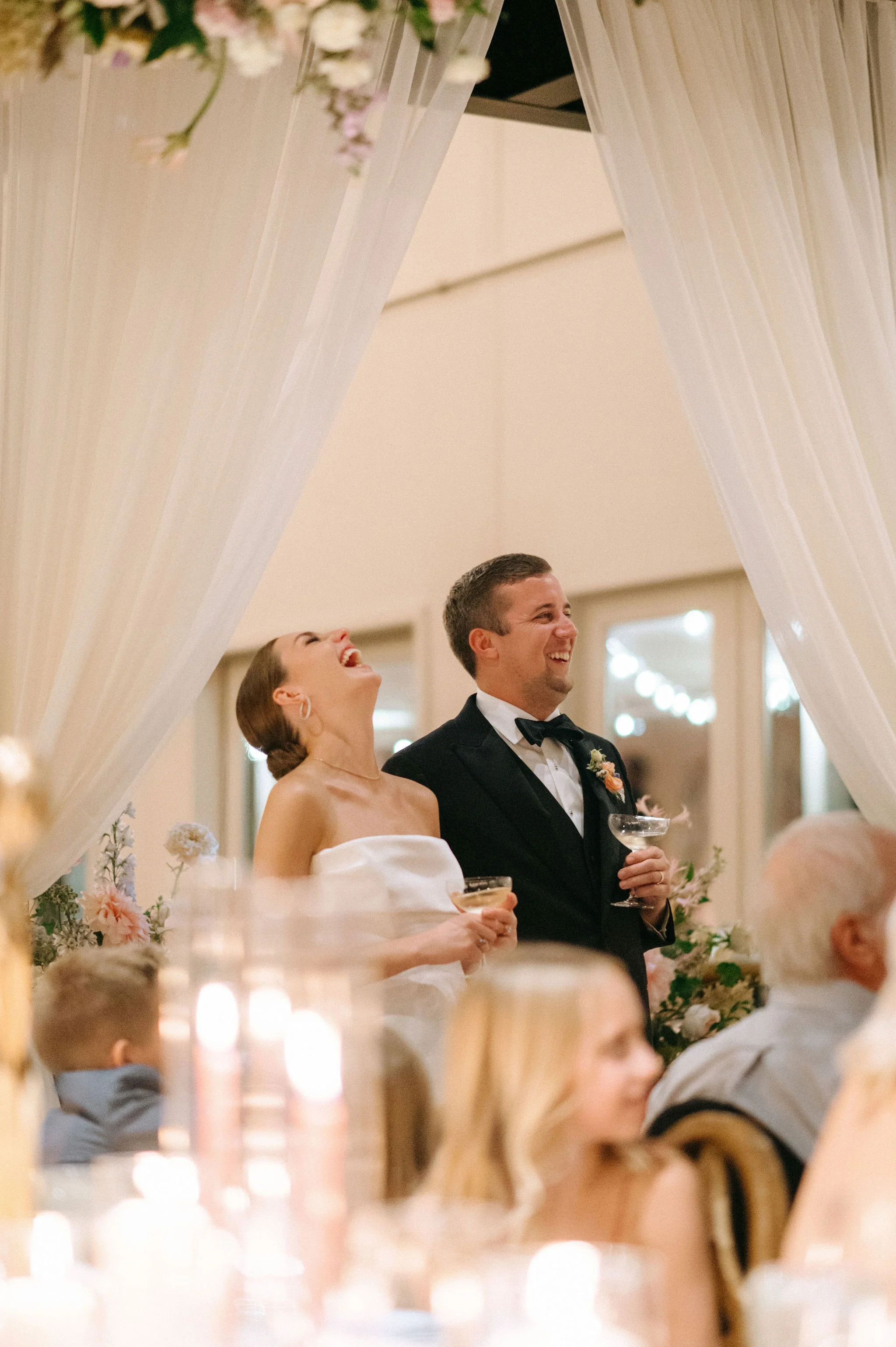 Cordelle bride and groom standing together at a wedding reception in the Victorian, smiling and holding drinks, surrounded by guests and floral decorations.