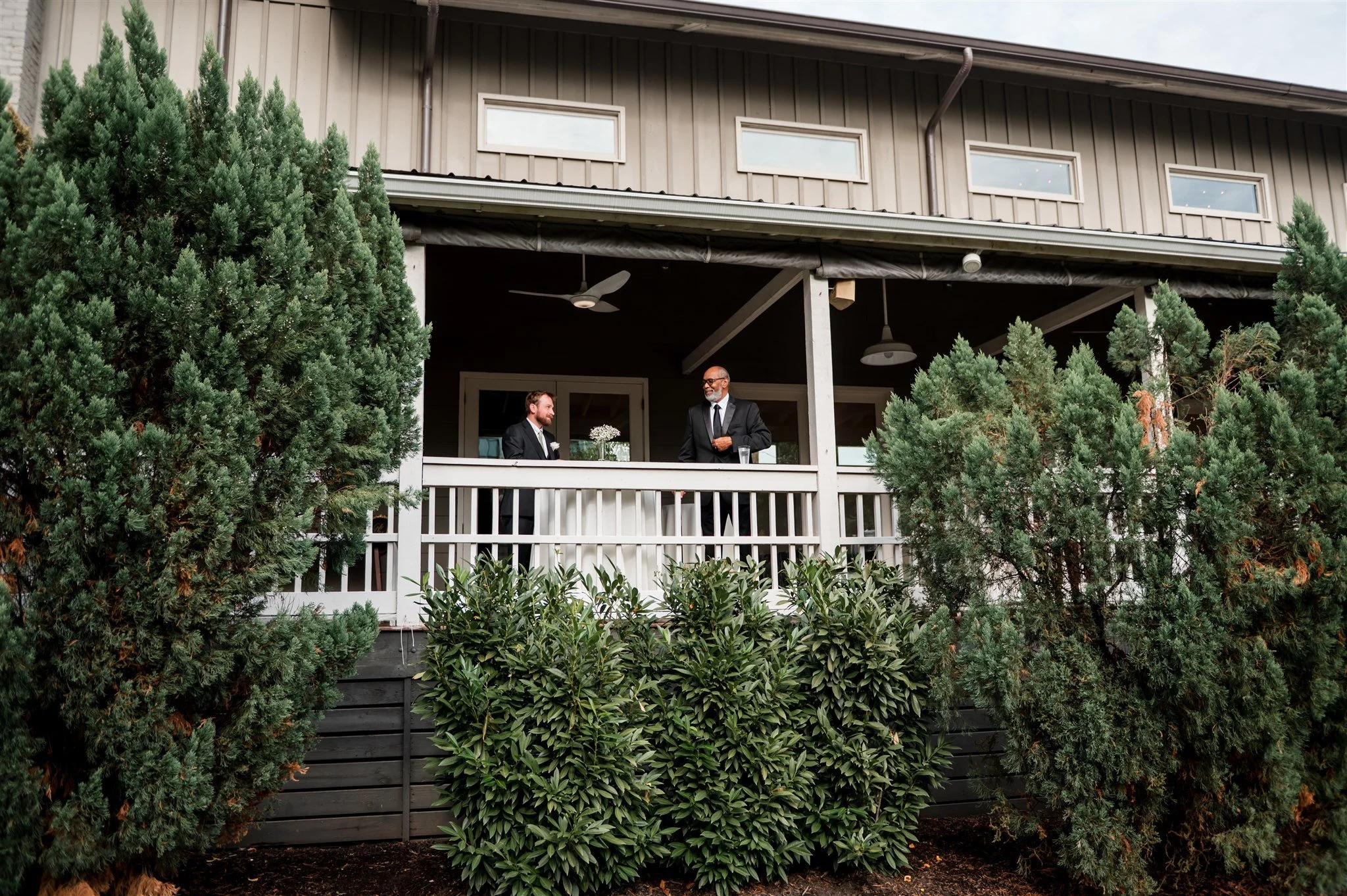 Two men dressed in suits are standing on the covered porch at the Cordlle, engaged in conversation. The scene is surrounded by lush green bushes and trees, with the house's upper windows visible above.