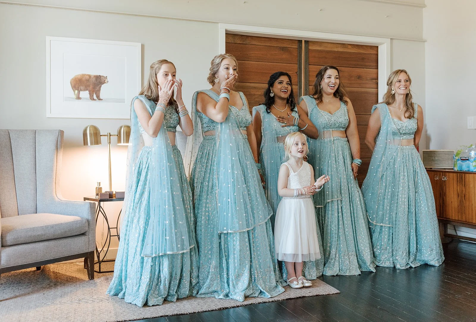 A bridal party in the loft dressed in matching light blue traditional dresses, standing in a living room with surprised and joyful expressions. Traditional bridal wear.