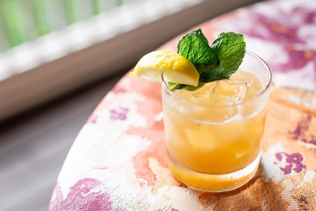 Signature cocktail for wedding reception in Nashville A glass of iced yellow cocktail garnished with a lemon wedge and fresh mint leaves on a colorful tablecloth.