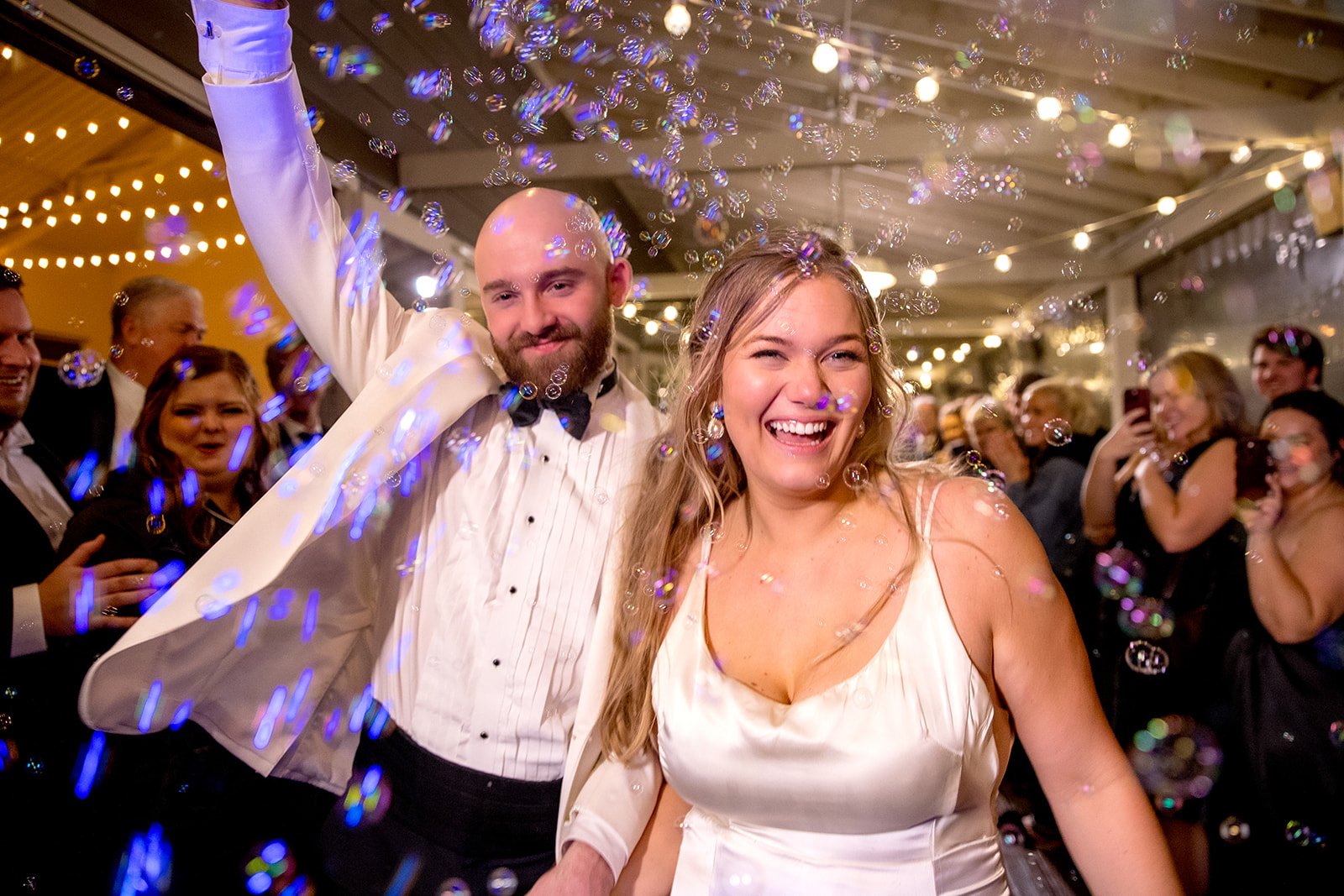 Wedding send-off at the Cordelle creating memories with guests Smiling couple celebrating at a wedding reception with guests and bubbles in the background.