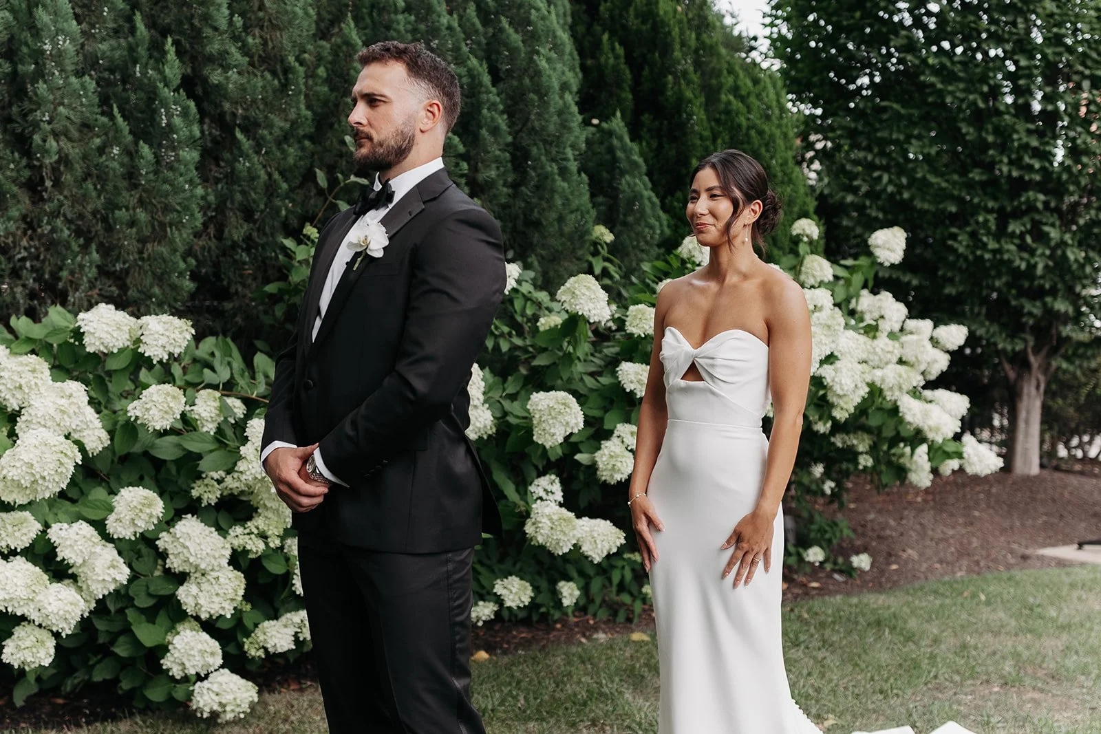 A Cordelle bride and groom standing outdoors during a wedding ceremony, with the groom in a black tuxedo and the bride in a white strapless wedding gown, amidst white hydrangea flowers and green foliage.