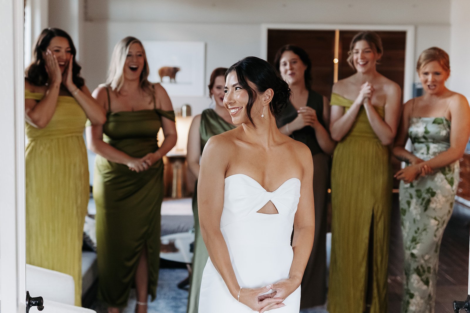 A bride in a white strapless wedding dress standing in front of a group of women, possibly her bridesmaids, who are reacting happily and emotionally to the moment in the loft at the Cordelle.