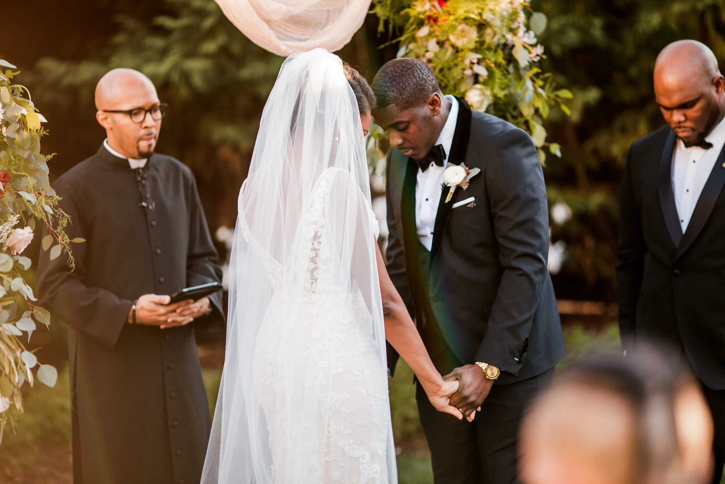A wedding ceremony in the Cordelle with a couple getting married in the garden with officiant and groomsmen, holding hands and bowing heads during the ceremony.