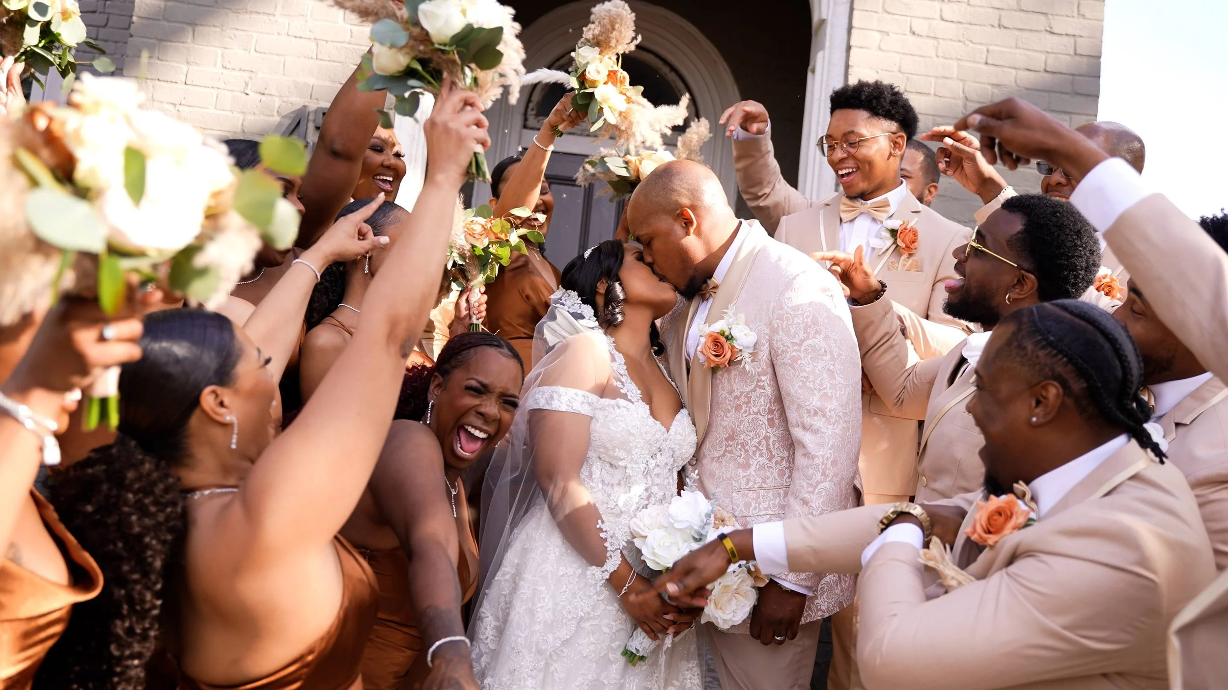 A wedding celebration with a Cordelle bride and groom kissing surrounded by friends and family holding bouquets and raising their hands in celebration outside a building.