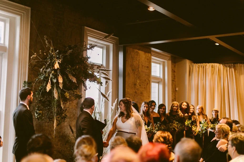 A Cordelle wedding ceremony in the Victorian with a bride and groom holding hands facing officiant, with bridesmaids in black dresses holding bouquets in background, inside a decorated venue.