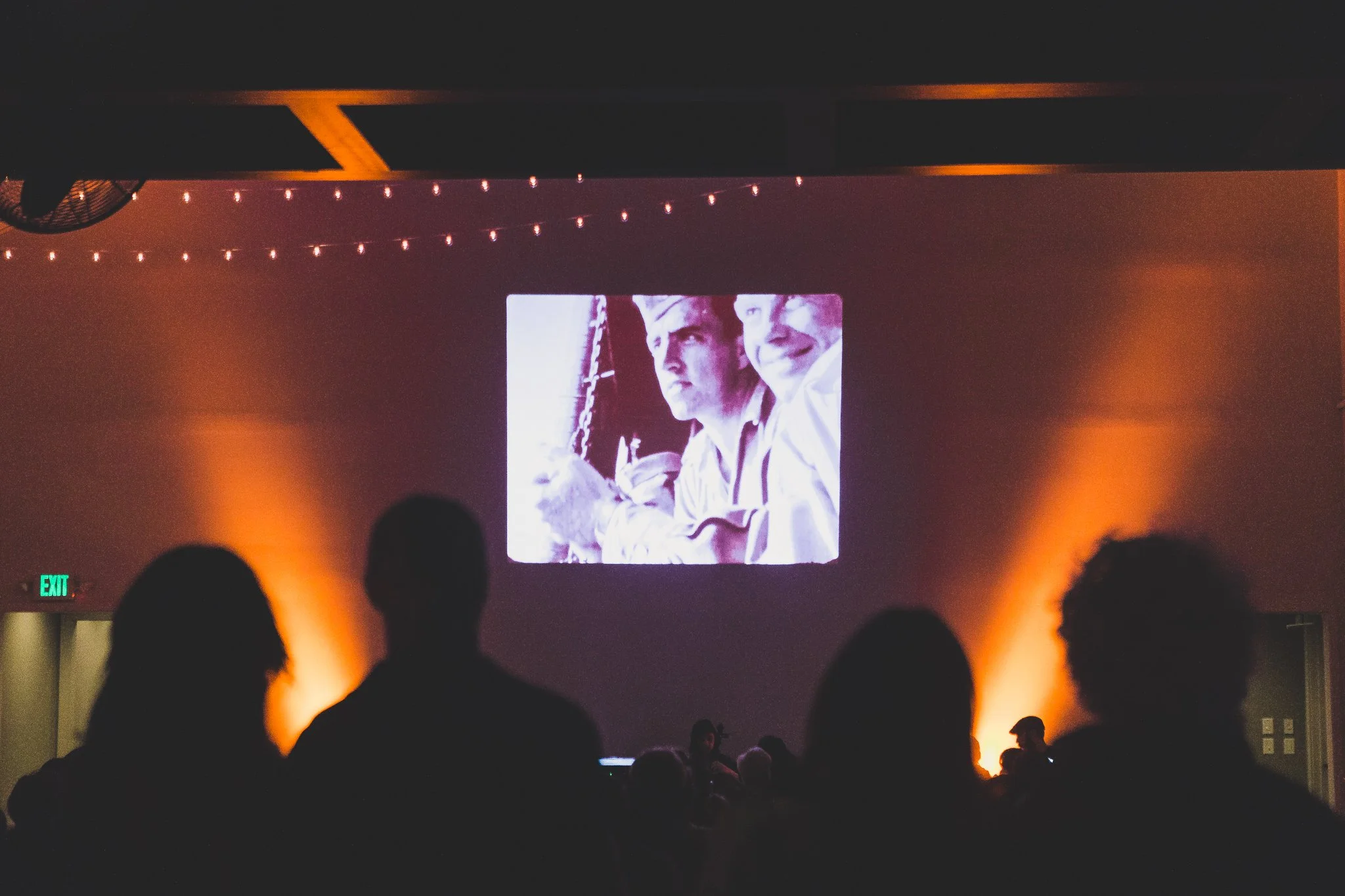 Movie night at the Cordelle in Nashville A dark indoor space with an audience facing a large screen displaying a black and white image of two men, one younger and one older, smiling.
