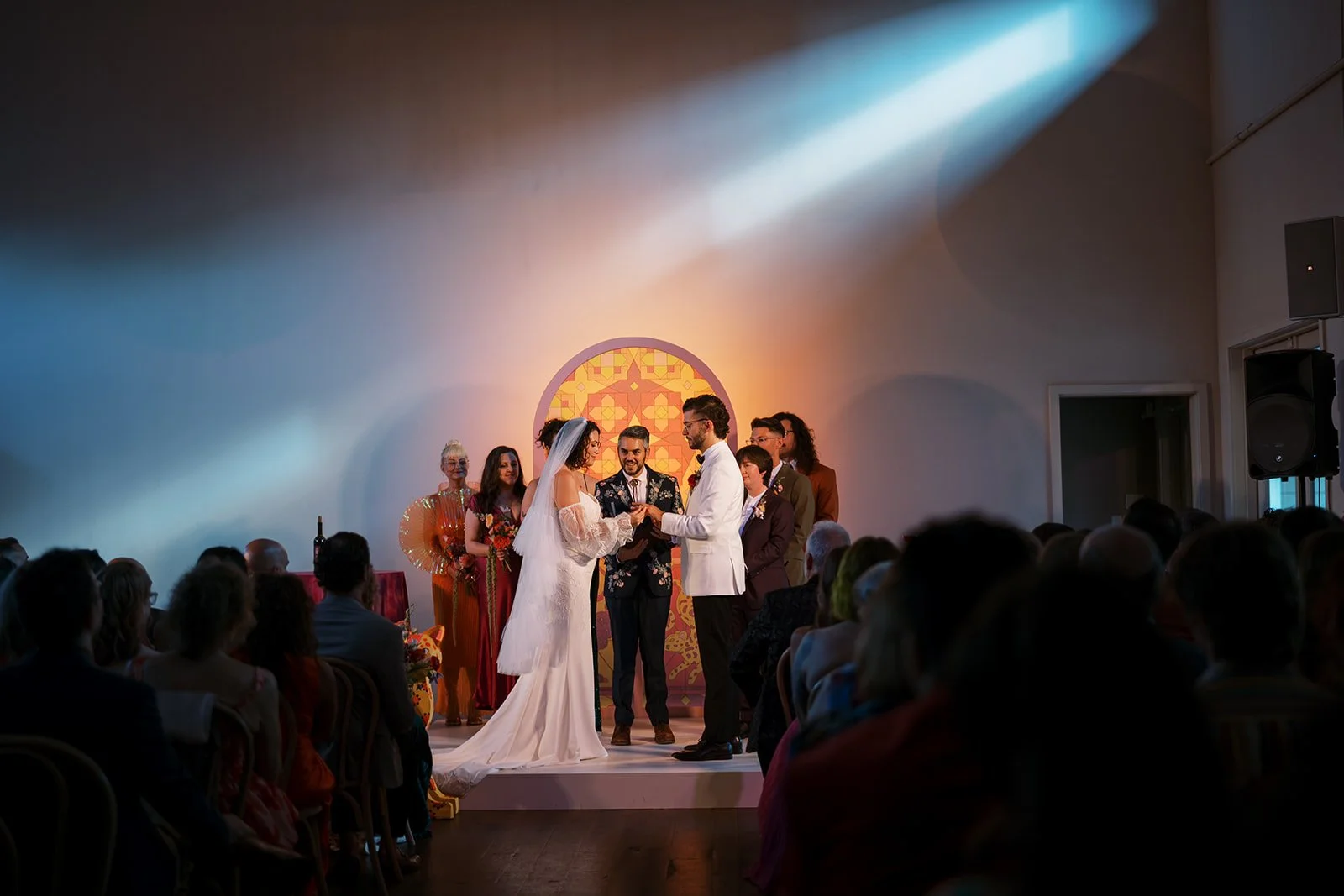 A wedding ceremony in the Great Hall at the Cordelle with a bride and groom exchanging rings on stage, surrounded by guests and officiant, with colorful decor and spotlights overhead.