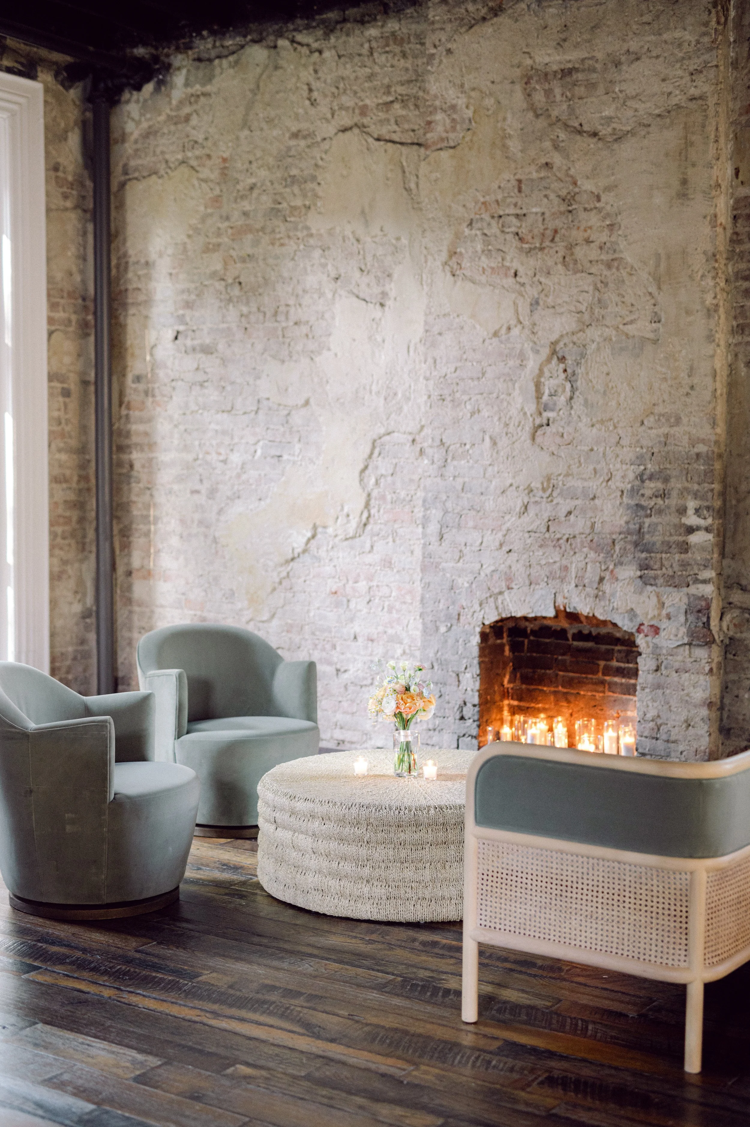 Lounge set rentals at the Cordelle cozy sitting area with a rustic exposed brick wall and a fireplace with candles. The space includes light gray armchairs, a round woven coffee table with a floral centerpiece, and a wooden floor with a dark finish.