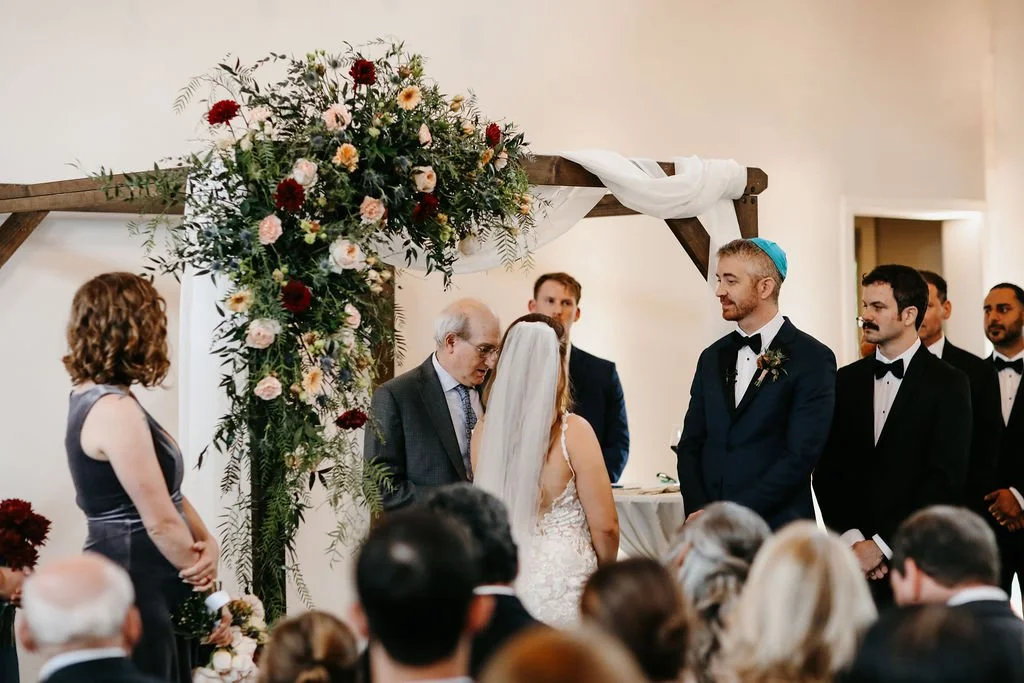 A Cordelle wedding ceremony in the Great Hall with a couple exchanging vows, surrounded by family and friends, under a floral arch decorated with flowers and white fabric.