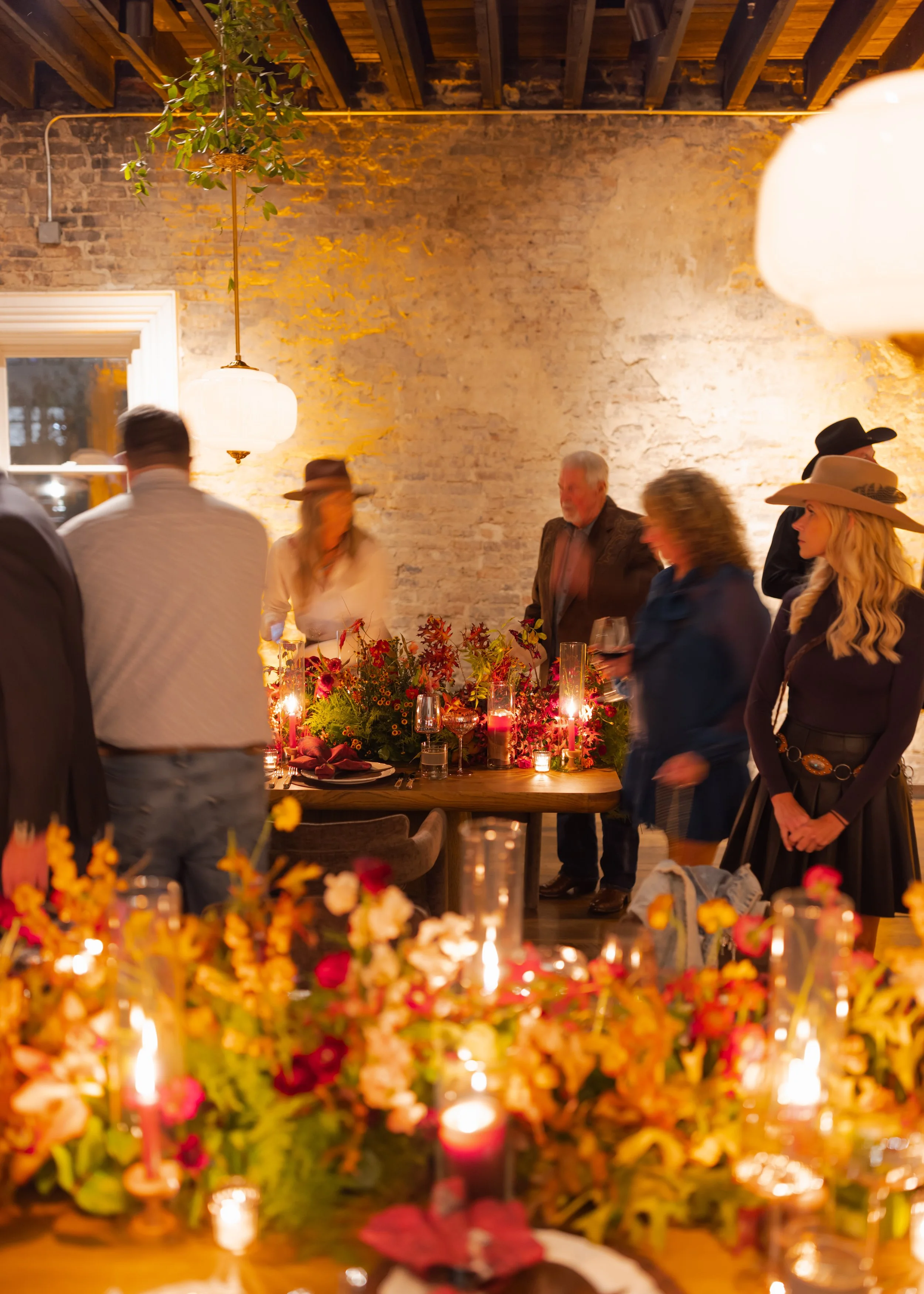 Corporate event at the Cordelle for CMA Awards People gathered around a decorated table with candles and flowers in a rustic indoor space with exposed brick and wooden ceiling beams.