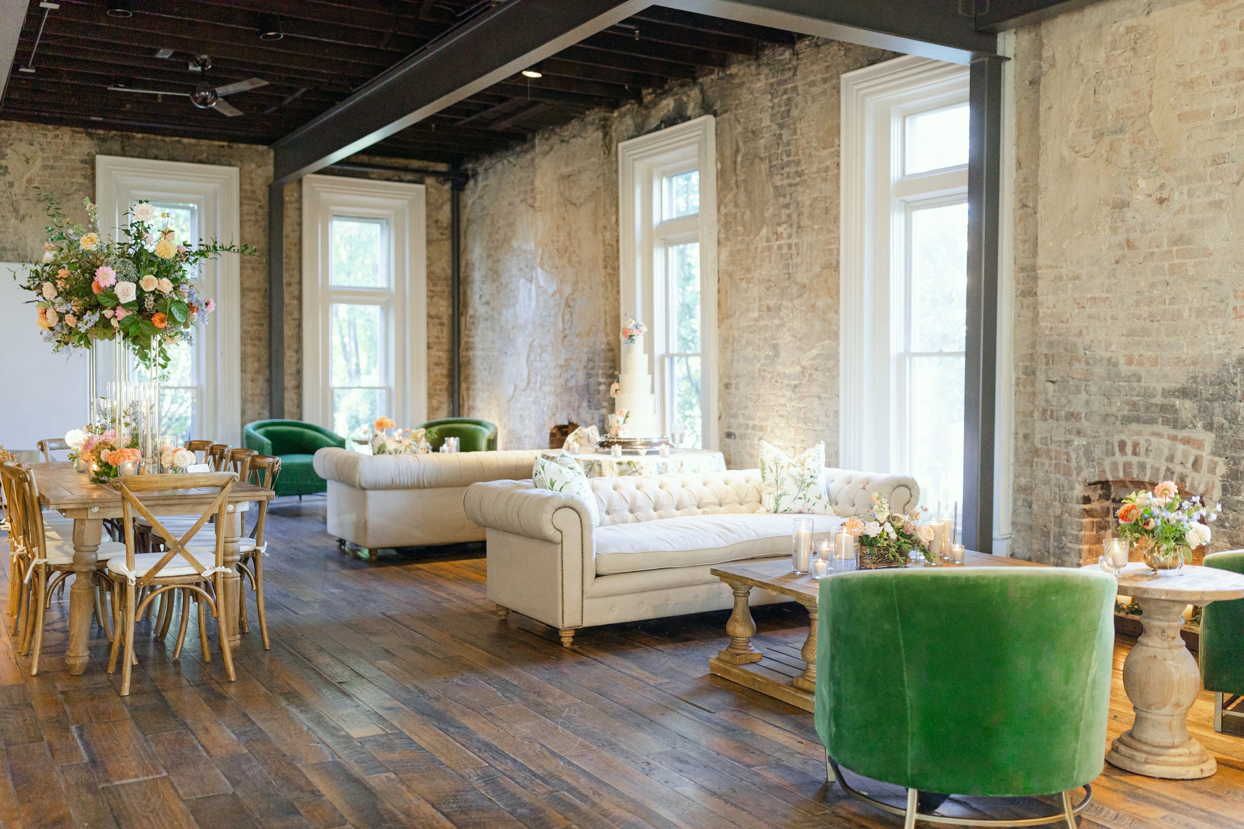 An interior room with exposed brick walls, large windows, a white tufted sofa, green velvet chairs, a wooden dining table with chairs, and floral centerpieces with candles on a wooden floor.