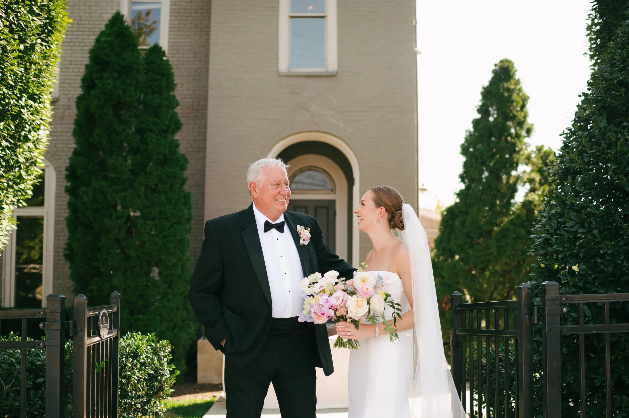 A bride in a white wedding dress holding a bouquet of flowers, smiling and looking at an her dad an older man in a black tuxedo with a bowtie, outside in front of a house with brick walls and green trees at the Cordelle Entrance in Nashville