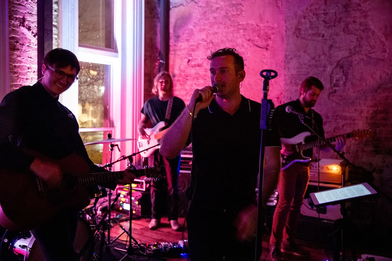 Record release at the Cordelle in the Victorian A musical band performing indoors with dark pink lighting, featuring a guitarist, a singer, and a bass player, against exposed brick walls and large windows.