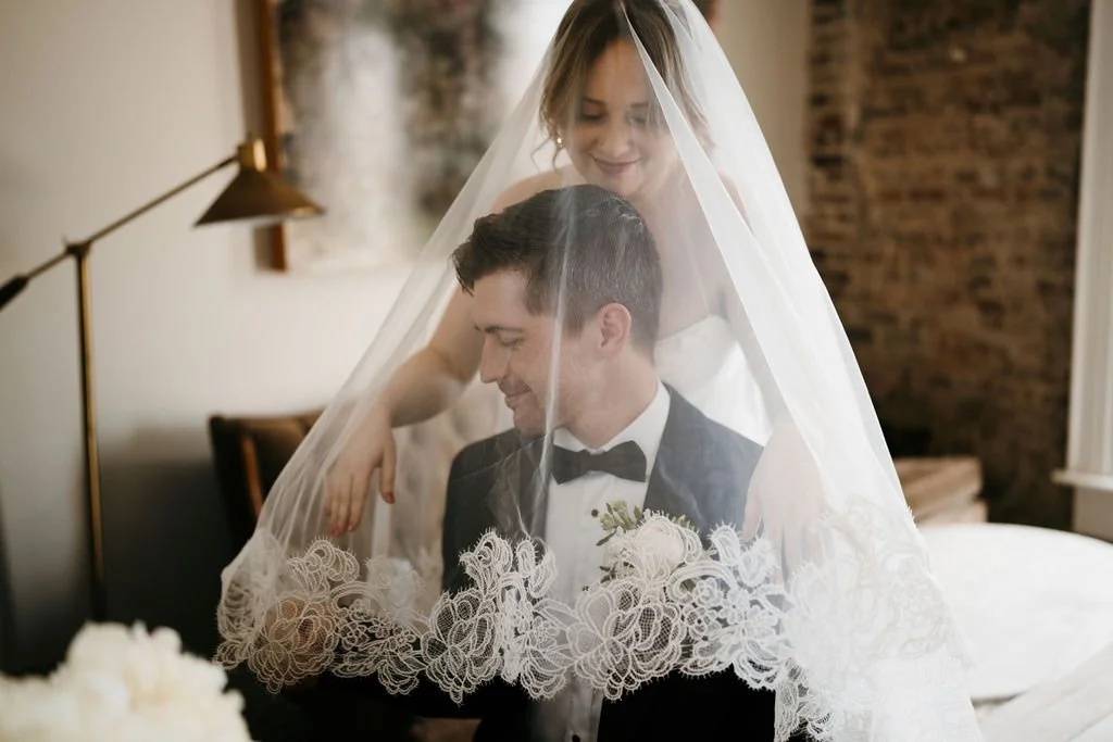 A bride and groom in the bridal suite at the Cordelle on their wedding day, with the bride standing behind and draping her veil over the groom's head, both smiling gently inside a cozy room.