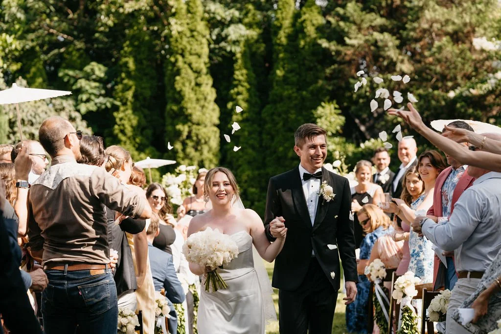 Bride and groom smiling and walking arm in arm through the Garden at the Cordelle guests during outdoor wedding celebration with floral aisle and greenery in background.