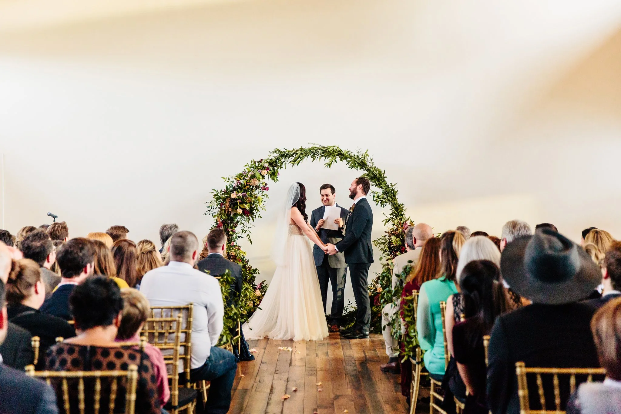 A wedding ceremony in the Great Hall at the Cordelle with a bride and groom holding hands under a floral arch, officiant reading vows, seated guests, in a large room with white walls and wooden floor. Modern event space