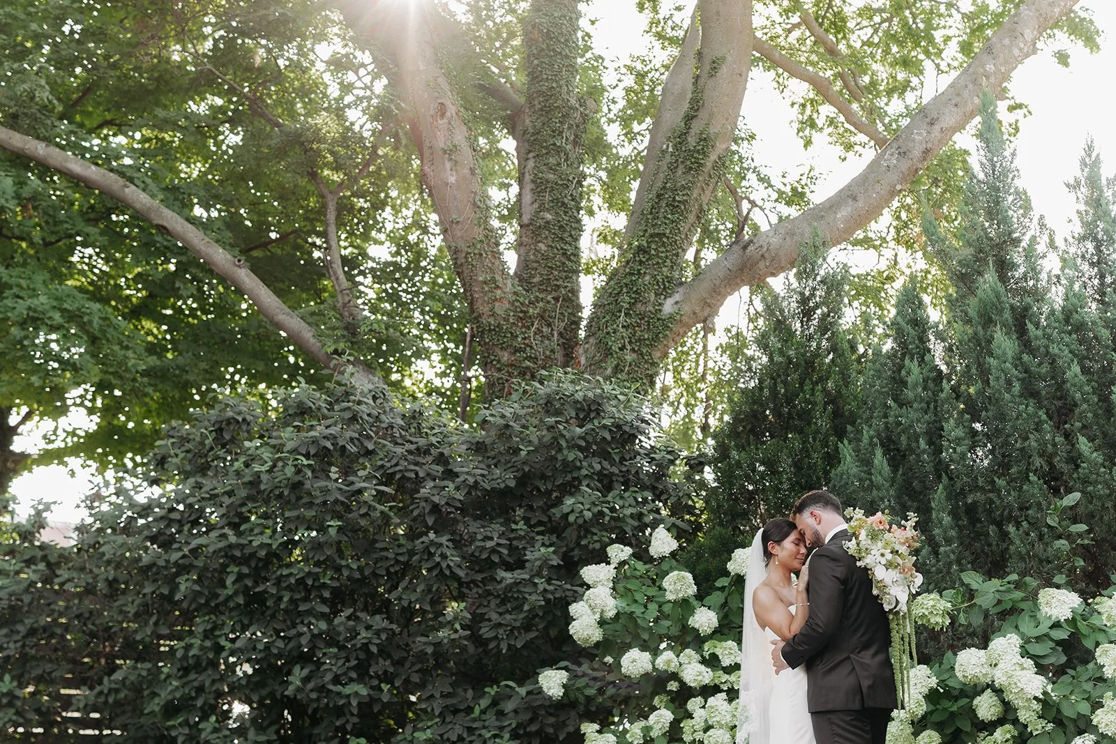 A bride and groom standing close together outdoors in a lush garden, surrounded by green trees and white flowering bushes, with the bride holding a large bouquet of flowers in the Garden in Downtown Nashville.