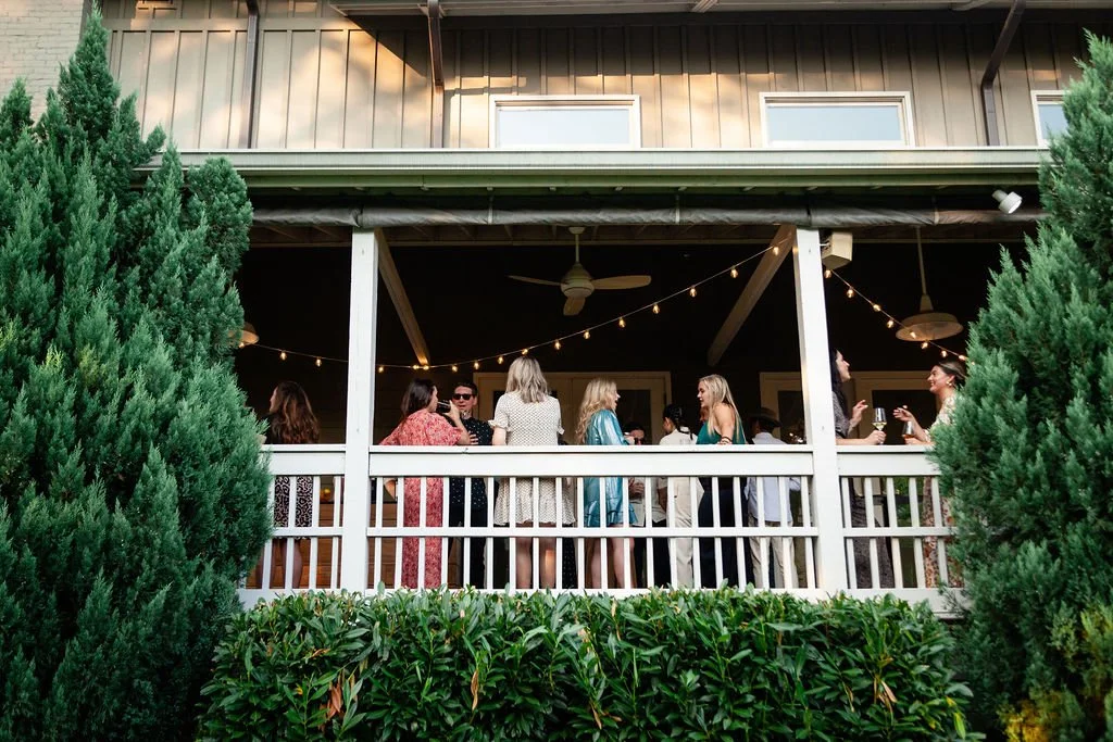 People socializing on a covered porch at the Cordelle with string lights, surrounded by green bushes and trees, during daylight.