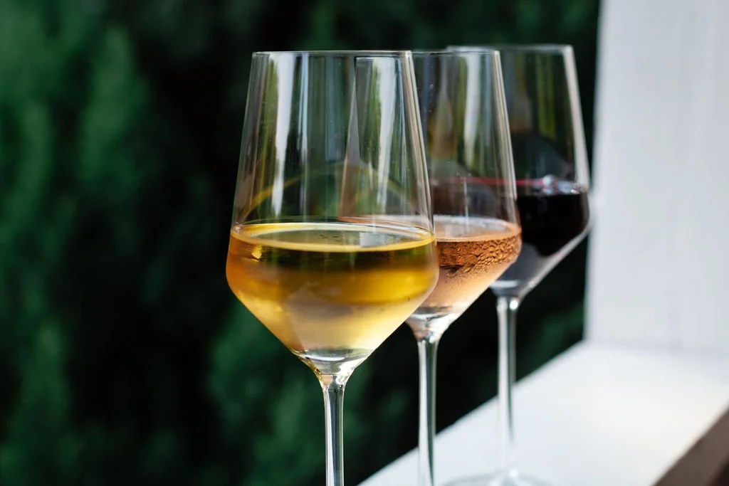 Three wine glasses filled with white wine, rosé, and red wine, placed on a white surface with a blurred green background.