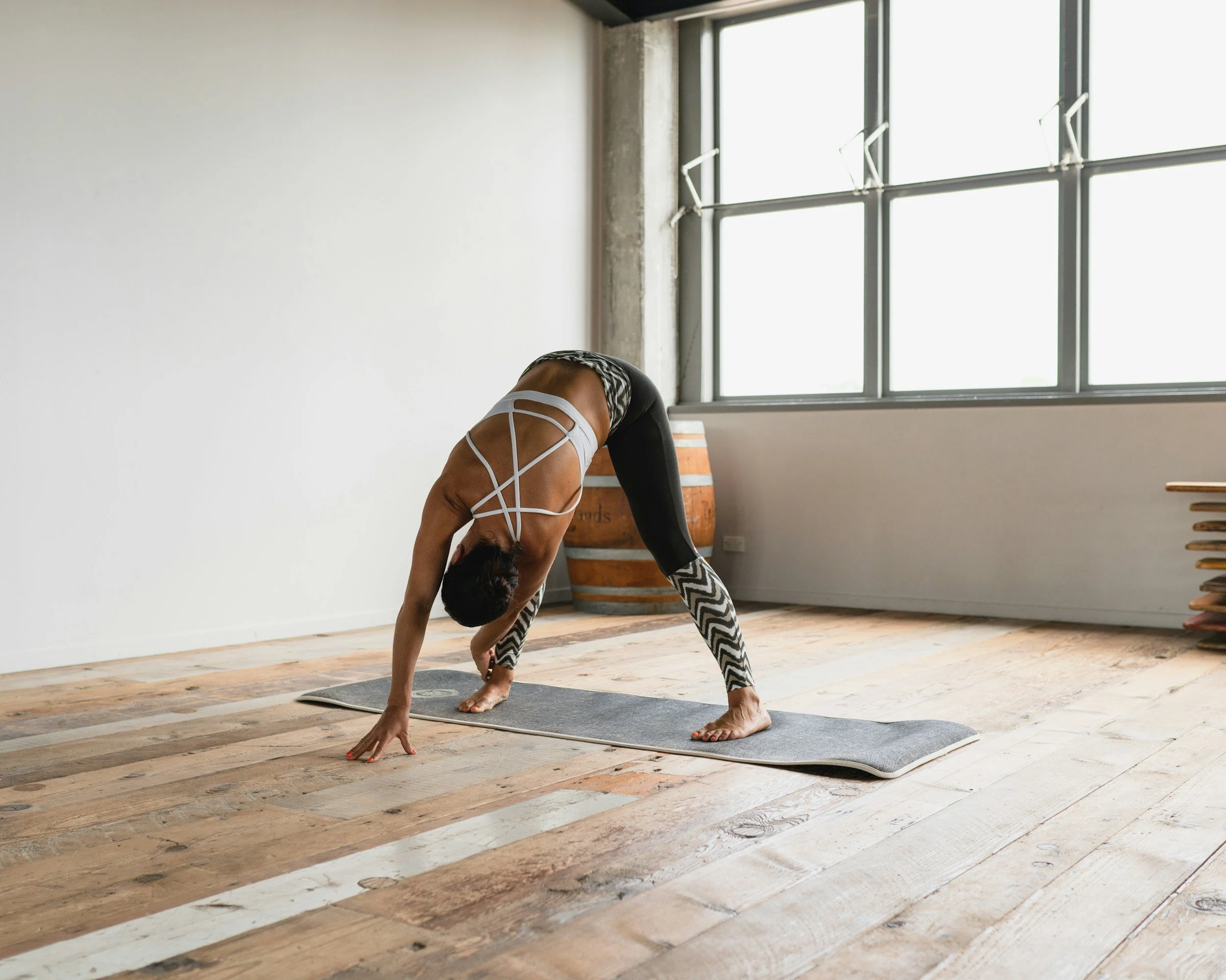 A woman practicing yoga in a spacious studio with large windows, wearing black leggings and a white strappy sports bra, positioned in a forward bend pose on a gray yoga mat.