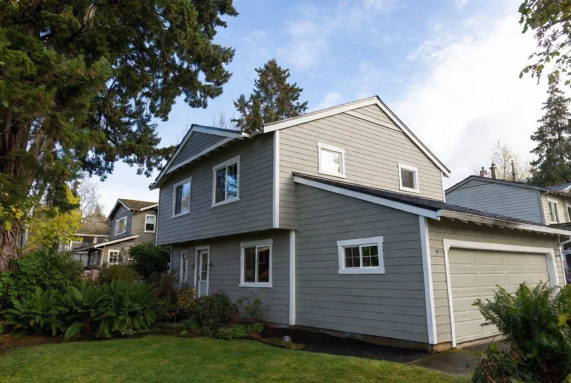 A two-story gray house with white trim, surrounded by green bushes and trees, with a partly cloudy sky overhead.