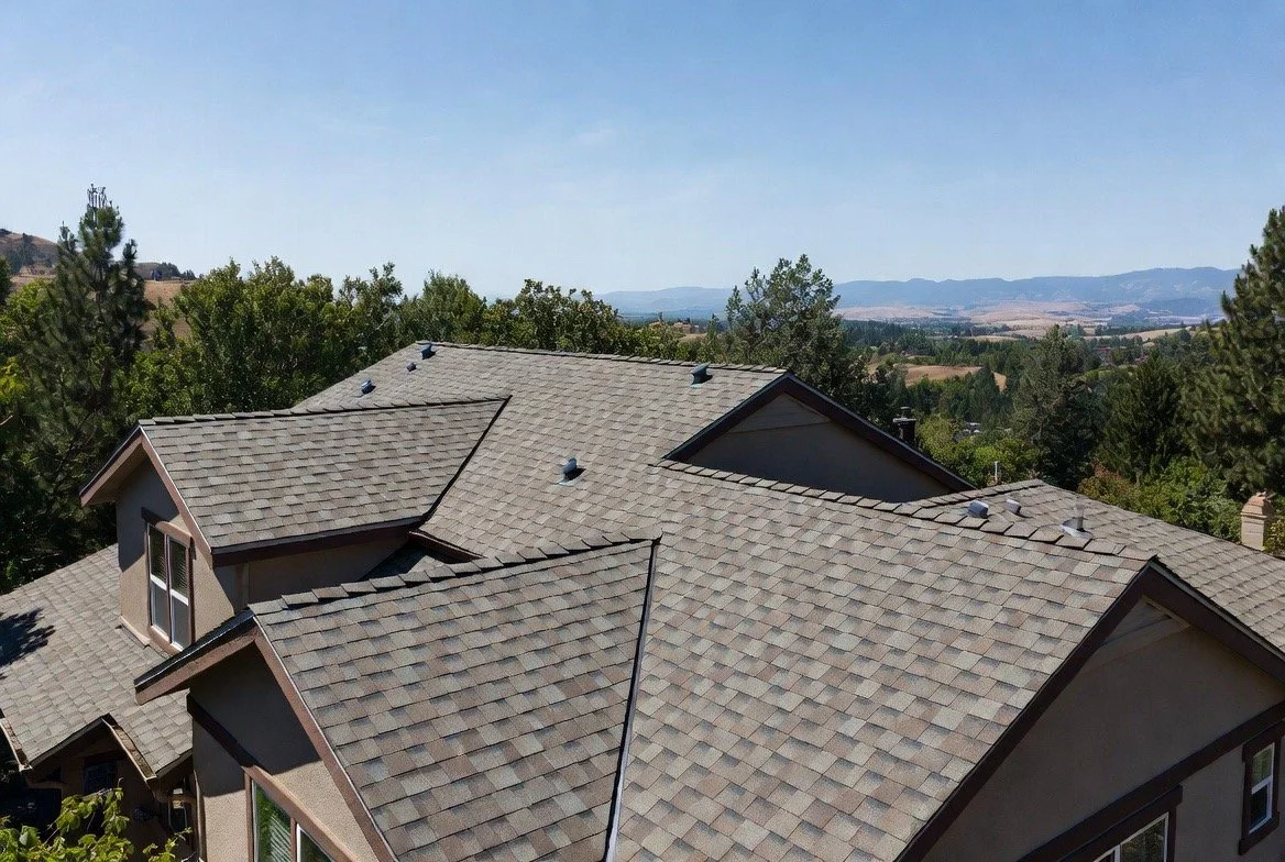A view of a house roof with asphalt shingles in a suburban area, surrounded by trees and distant hills under a blue sky.