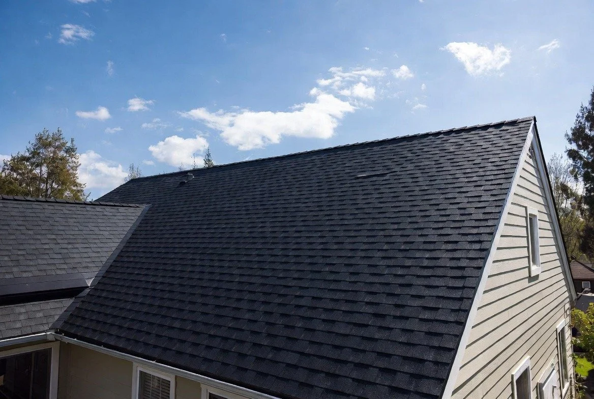 View of a house roof with gray shingles, under a blue sky with some clouds.