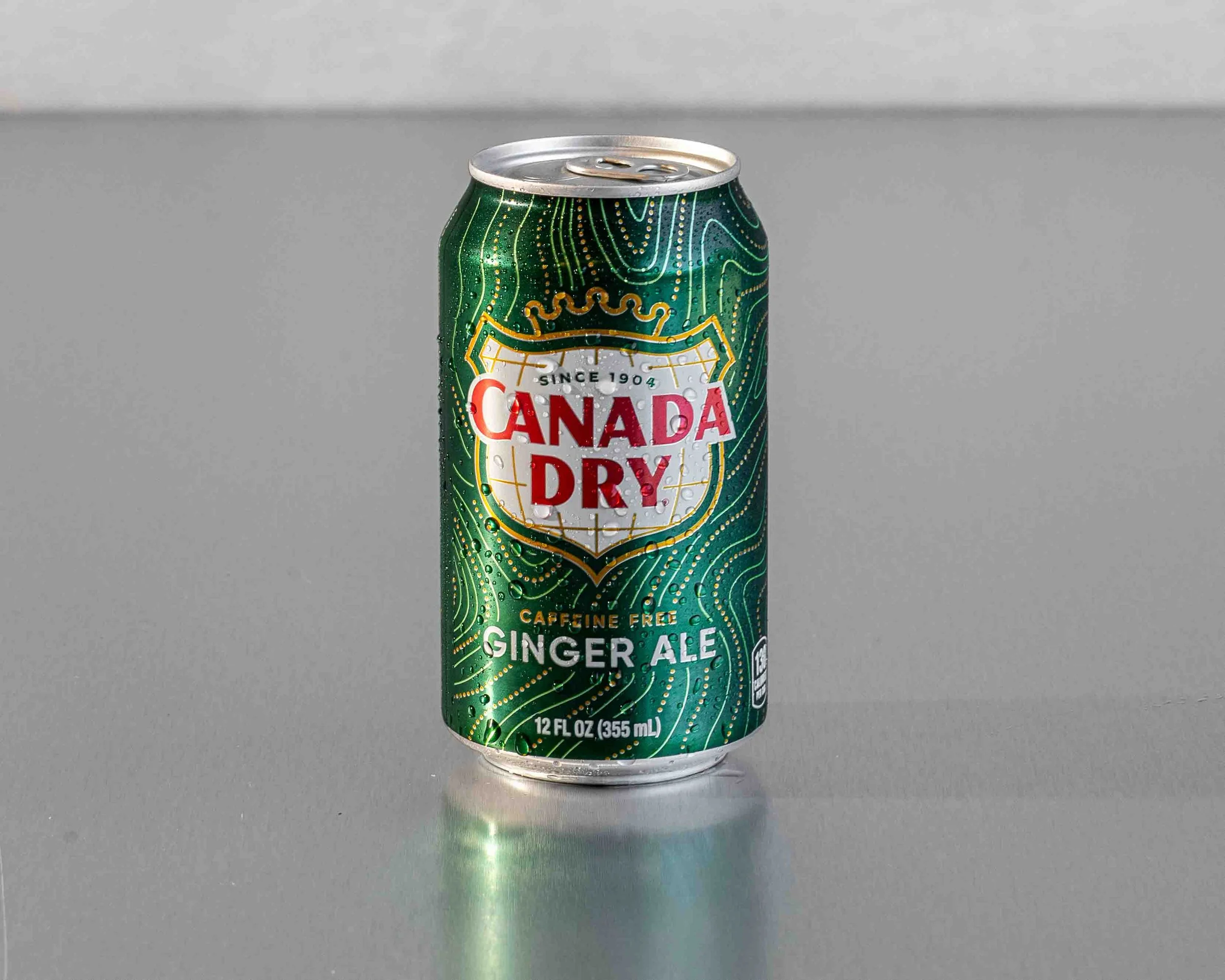 A can of Canada Dry Ginger Ale with green and gold design, labeled as caffeine free, standing on a flat surface with a plain, light-colored background.