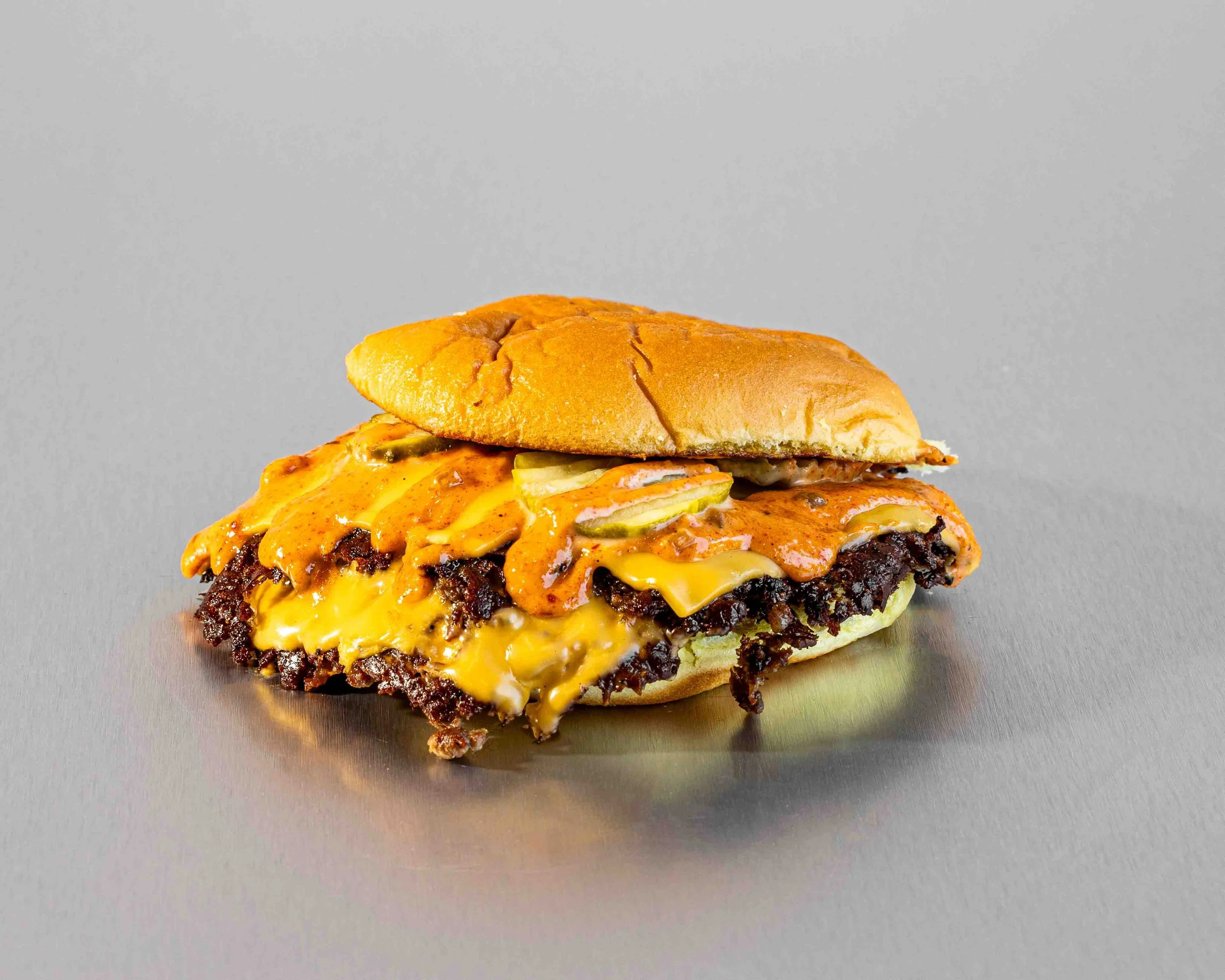 Close-up of a cheeseburger with melted cheese, pickles, and a toasted bun on a gray surface with a plain background.