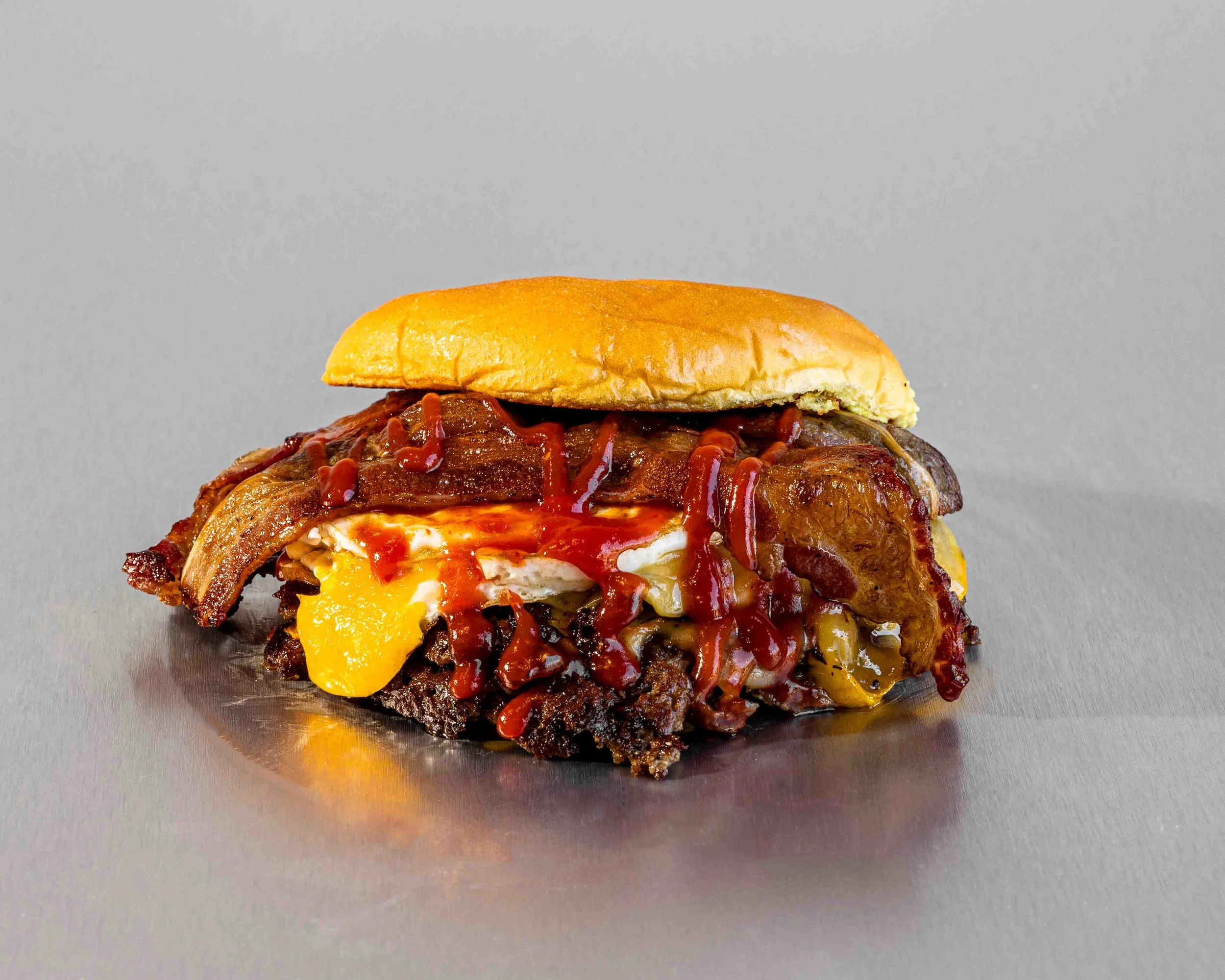 A close-up of a burger with bacon, cheese, and ketchup on a gray background.