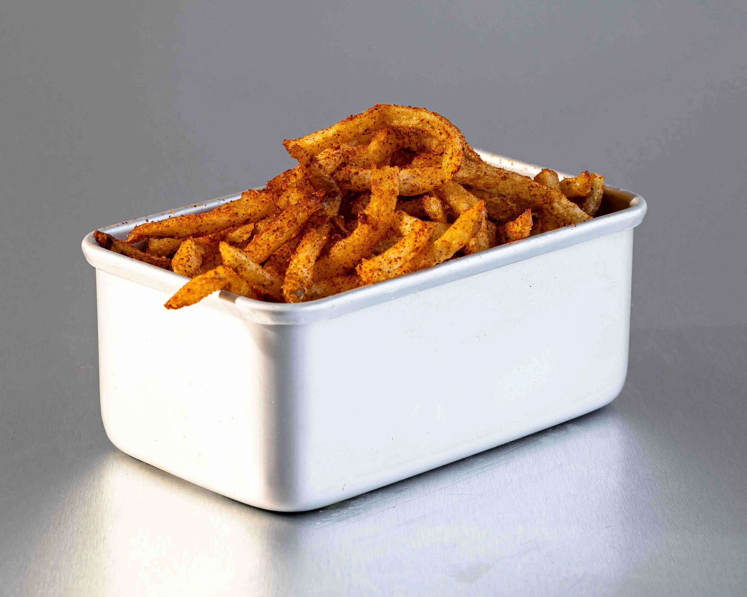 A white rectangular container filled with seasoned French fries, some of which are curled, on a gray surface with a plain gray background.