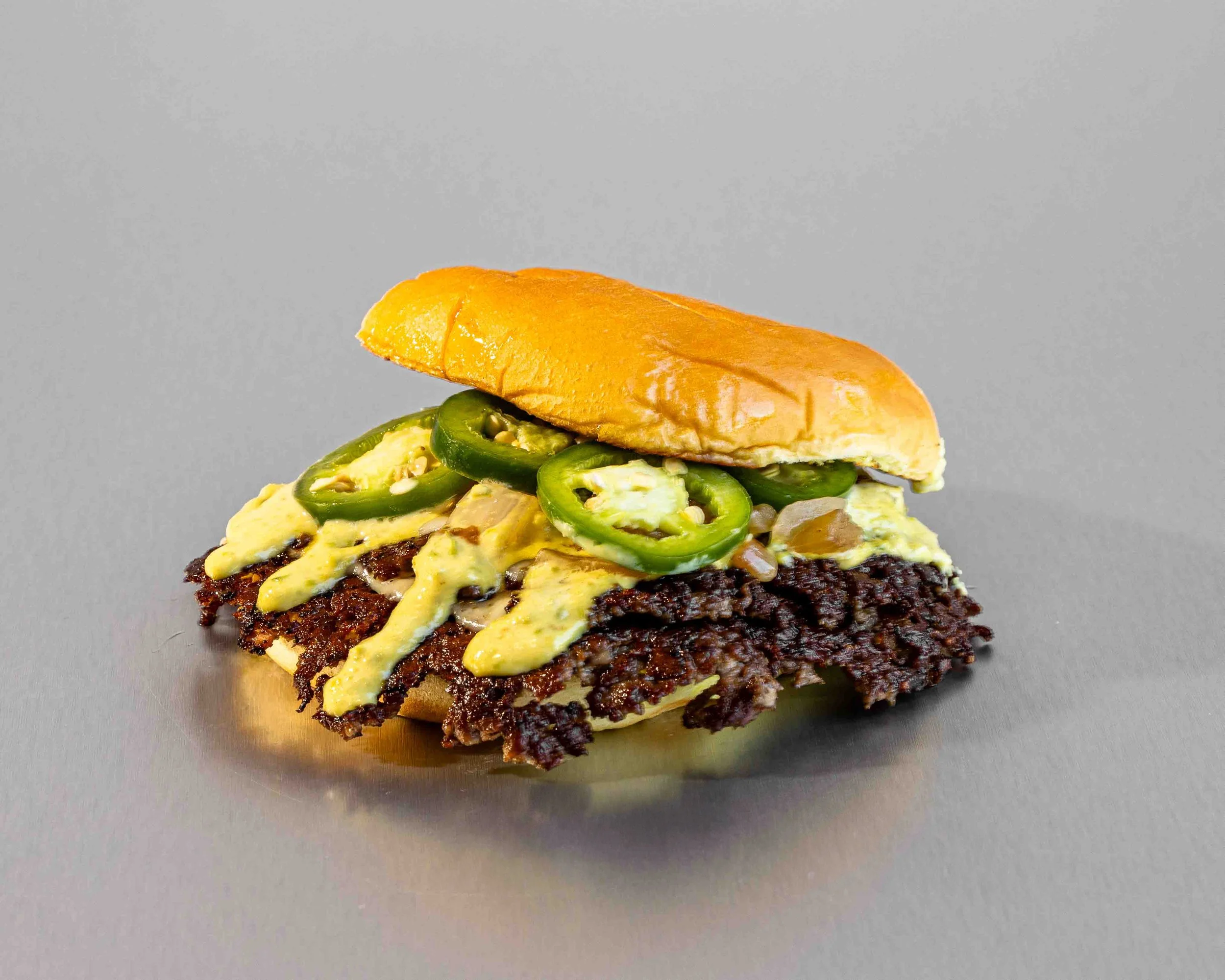 Close-up of a burger with jalapeño slices, melted cheese, and a glossy bun on a gray background.