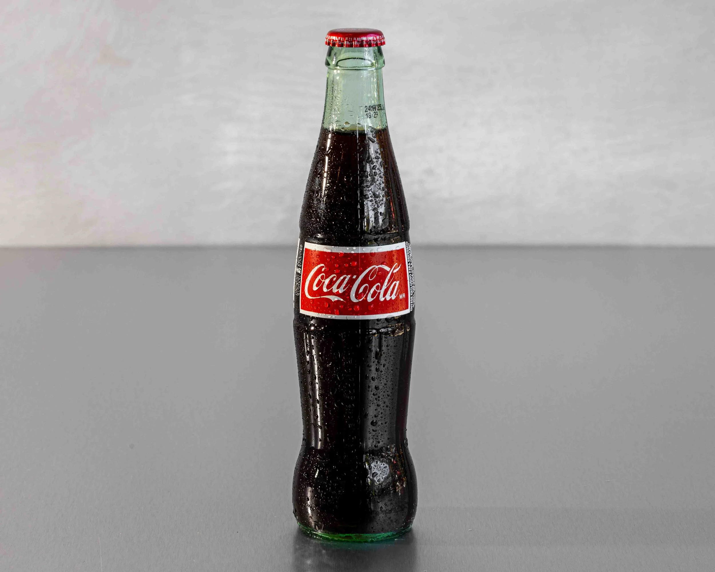 A glass Coca-Cola bottle with condensation droplets, positioned upright against a plain light gray background.