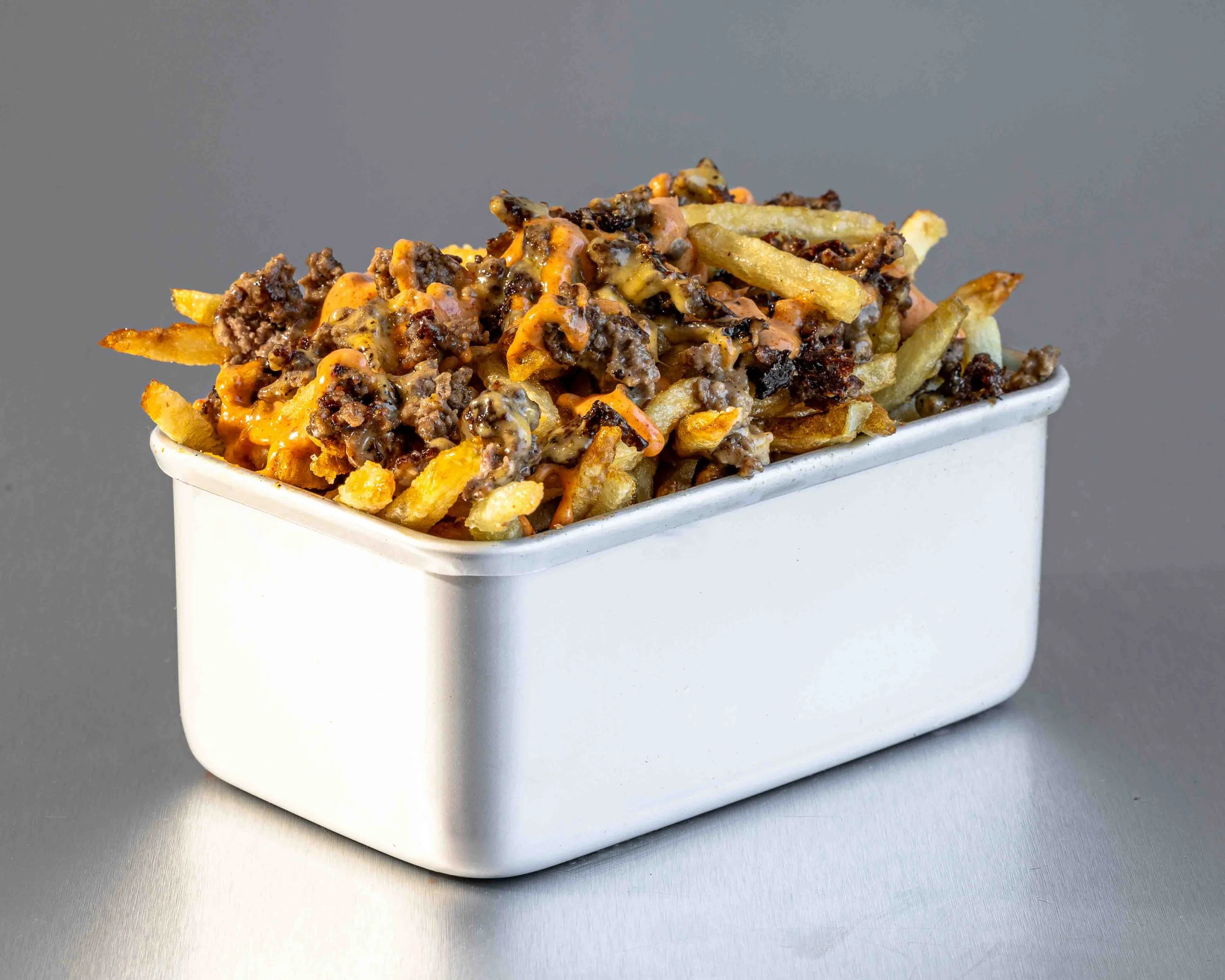A white rectangular container filled with loaded French fries topped with ground beef, melted cheese, and melted cheddar cheese, set against a gray background.
