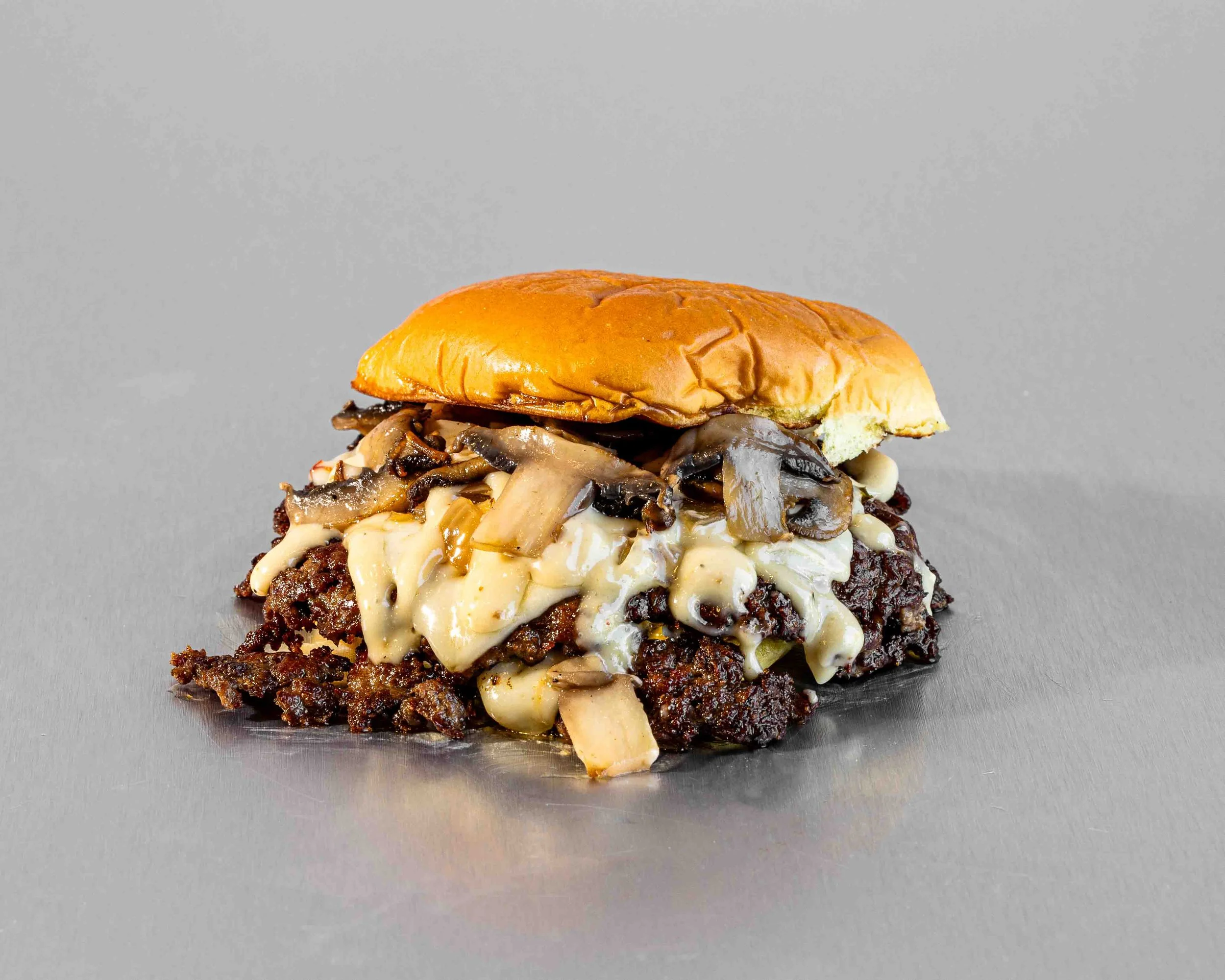 Close-up of a gourmet burger with sautéed mushrooms, melted cheese, and a toasted bun, placed on a metallic surface.
