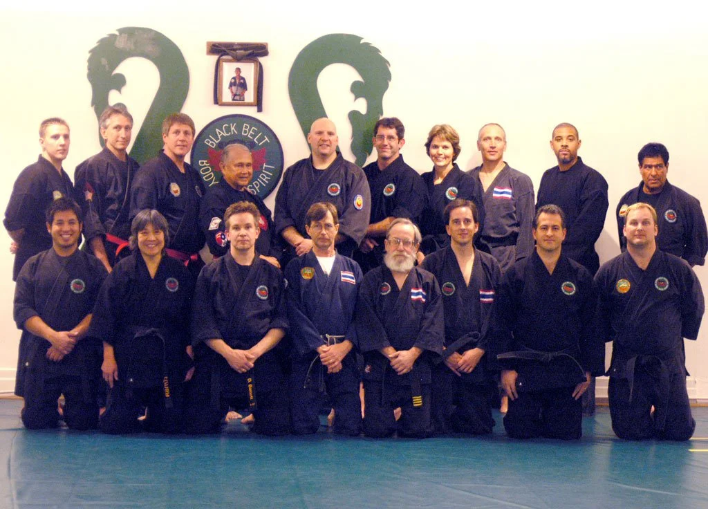 Group of martial arts students and instructors in black uniforms with patches, kneeling and standing on a mat inside a dojo, with a wall display featuring two dragon head silhouettes and a black belt with a patch.