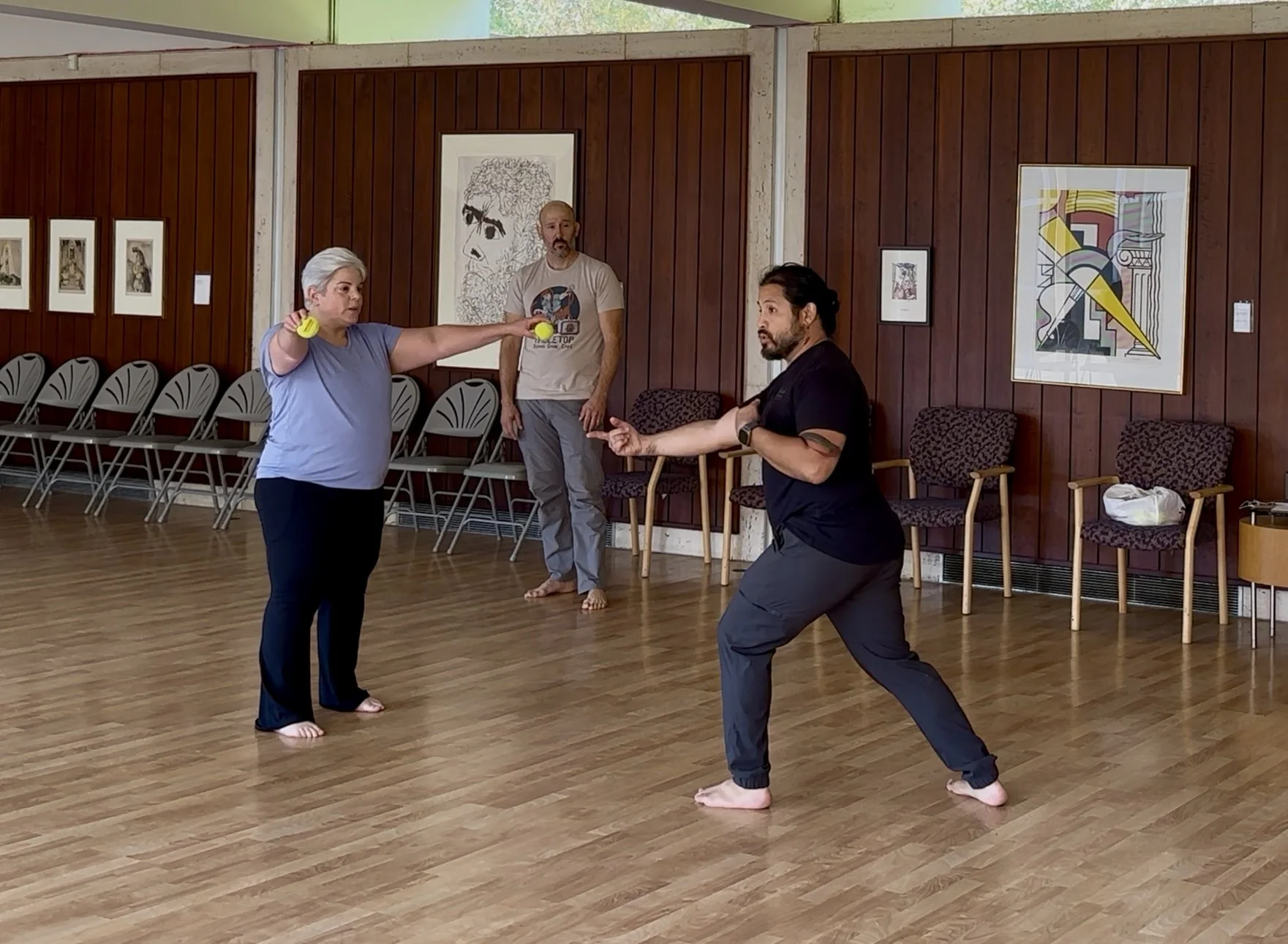 Three people practicing movement exercises, with wooden floors and art on the wall. One woman with gray hair holds yellow balls, a man crouches in a martial stance, and another man watches.