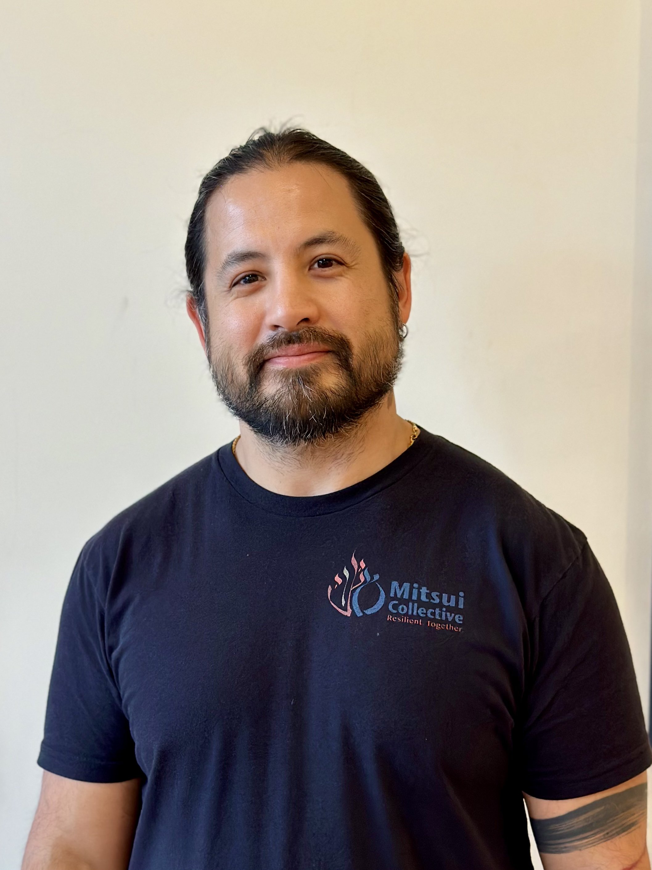 Close-up of a man with dark hair and beard, wearing a black T-shirt with a colorful logo and the words "Mitsui Collective" and "Resilient Together" on it, standing against a plain beige wall.