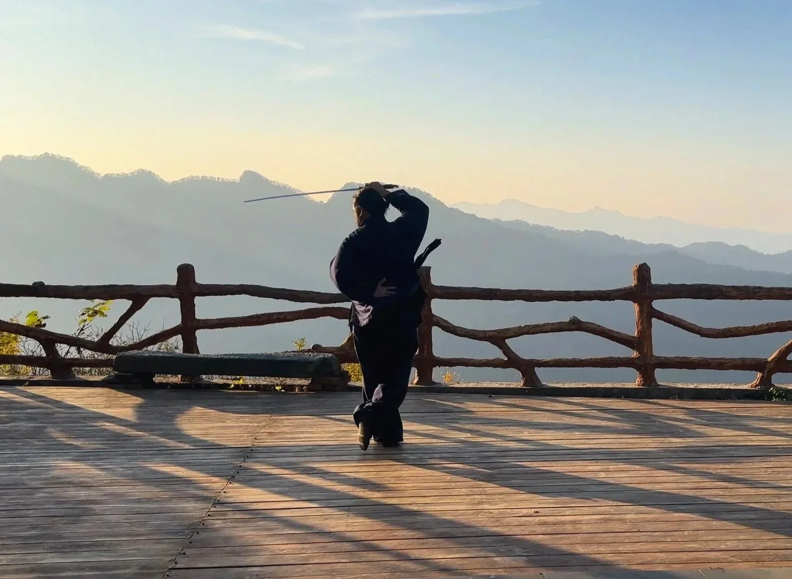 Person practicing martial arts on a wooden deck with mountain views and a rustic railing at sunset.