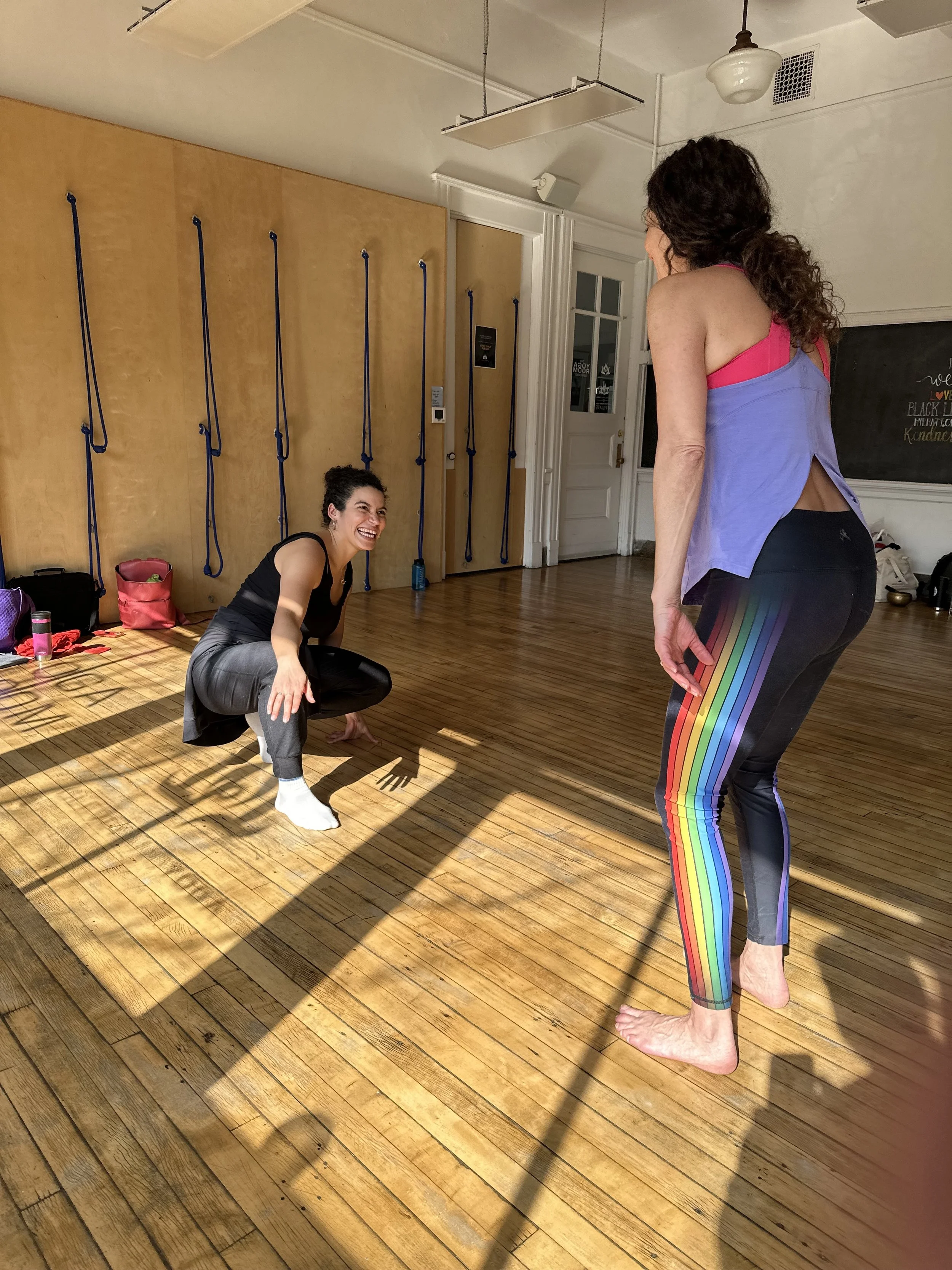 Two women are in a room with wooden floors, engaging in a friendly conversation. One woman is squatting and smiling, while the other woman, standing barefoot, is talking to her. The standing woman is dressed in rainbow-striped leggings and a pink and purple top. The room has a blackboard with colorful writing on it, a door, and a wall with yoga ropes.