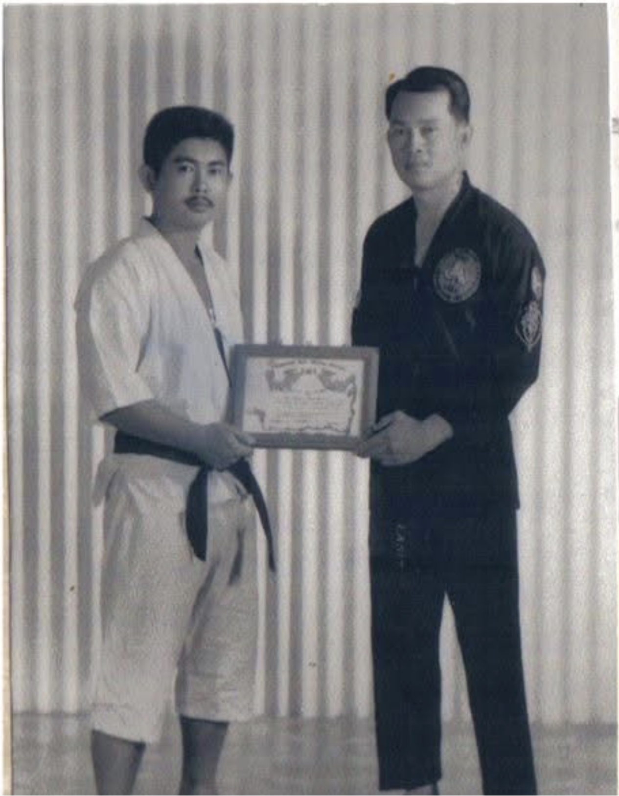 A woman in a karate uniform with a black belt receiving an award or certificate from a man in a martial arts uniform with patches, standing in front of a curtain.
