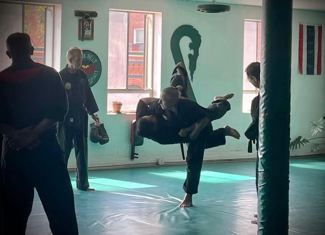 Martial arts class where an instructor demonstrates a throw on a student holding a pad, with four individuals observing in a martial arts studio, decorated with martial arts banners and flags.