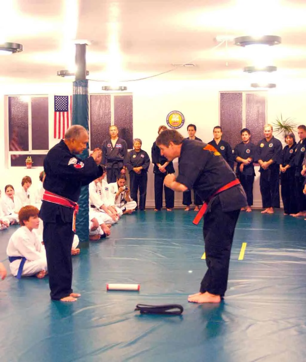 Martial arts class in a dojo with children and adults in uniform, two instructors bowing to each other in the center, with students seated on the floor and standing in the background.